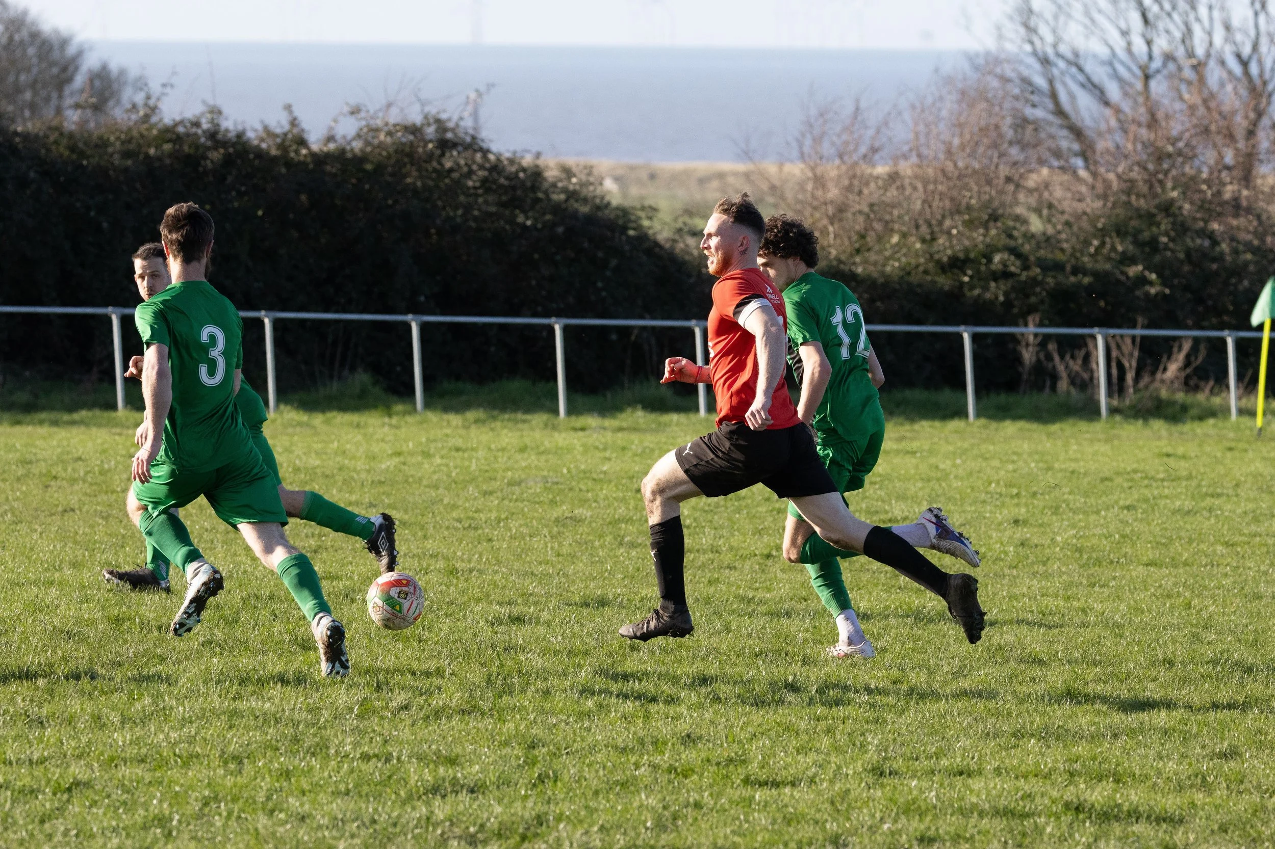 Soccer players actively running on a grassy field during a game, with some wearing green uniforms and one in a red uniform, against a backdrop of trees and open sky.