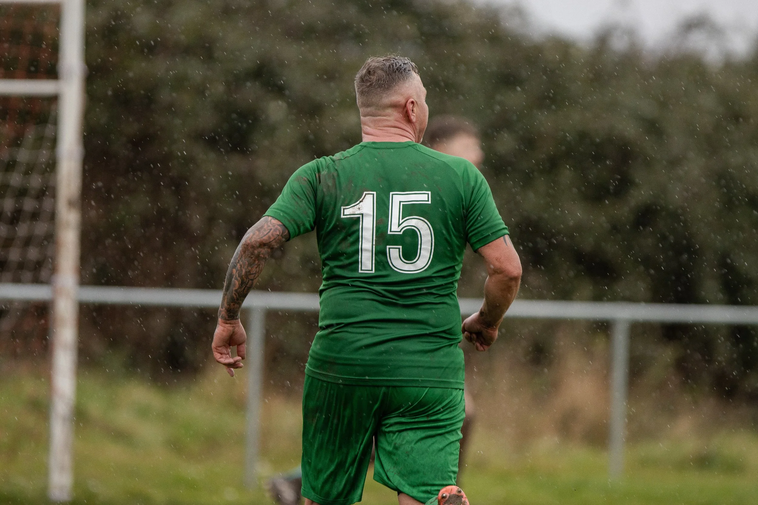 A male soccer player is on the field with his back to the camera, wearing a green jersey with the number 15 and matching shorts, during a rainy match.