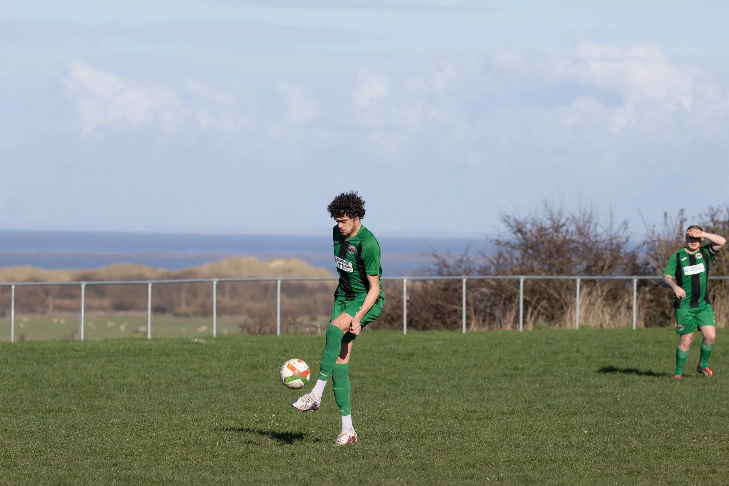 A soccer player in a green uniform stands on a grass field, preparing to kick a soccer ball, with another player in similar attire in the background shielding his eyes from the sun.