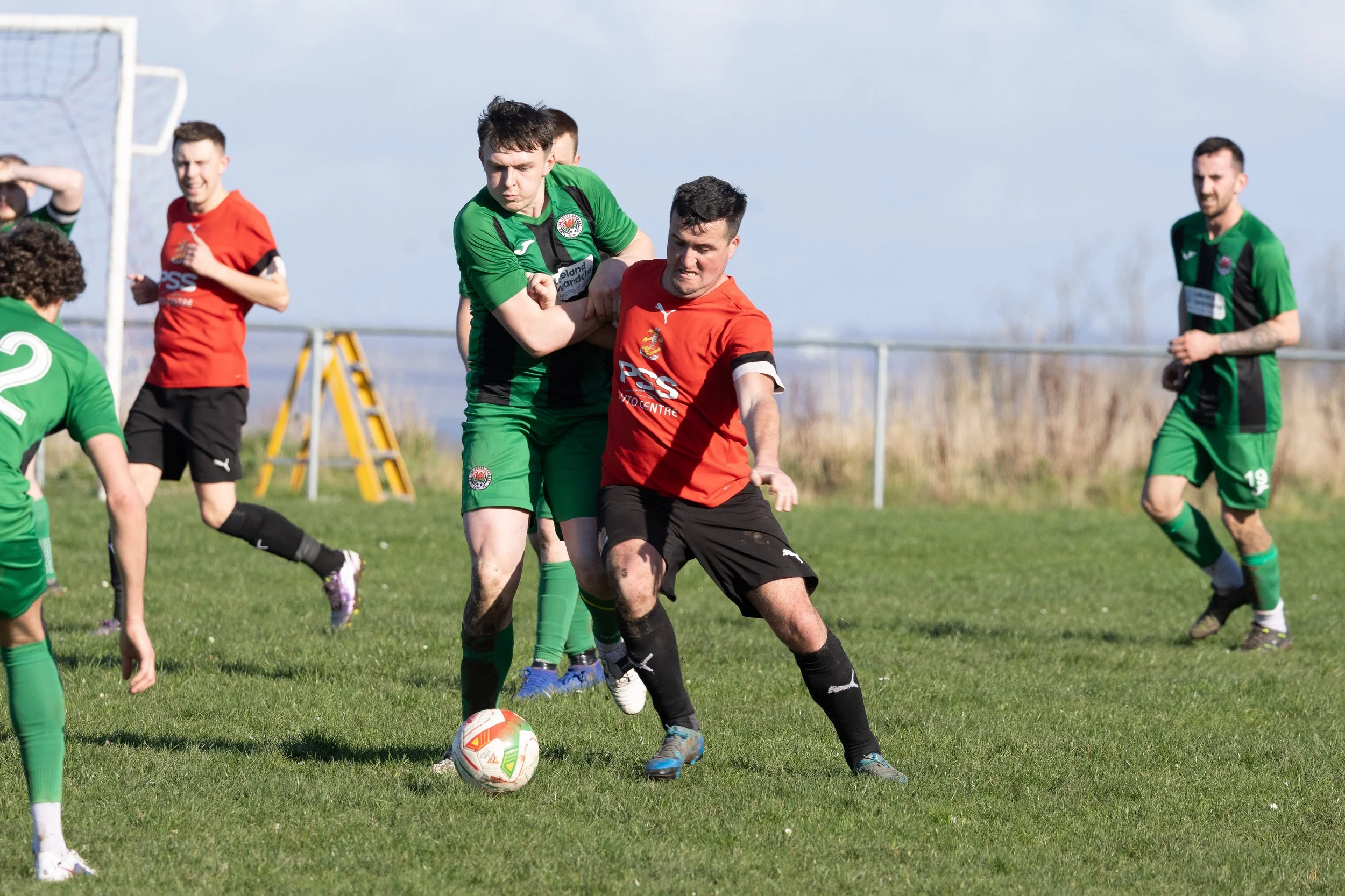 Soccer players in green and red jerseys competing on a grassy field, with some players in background and a goalpost visible.