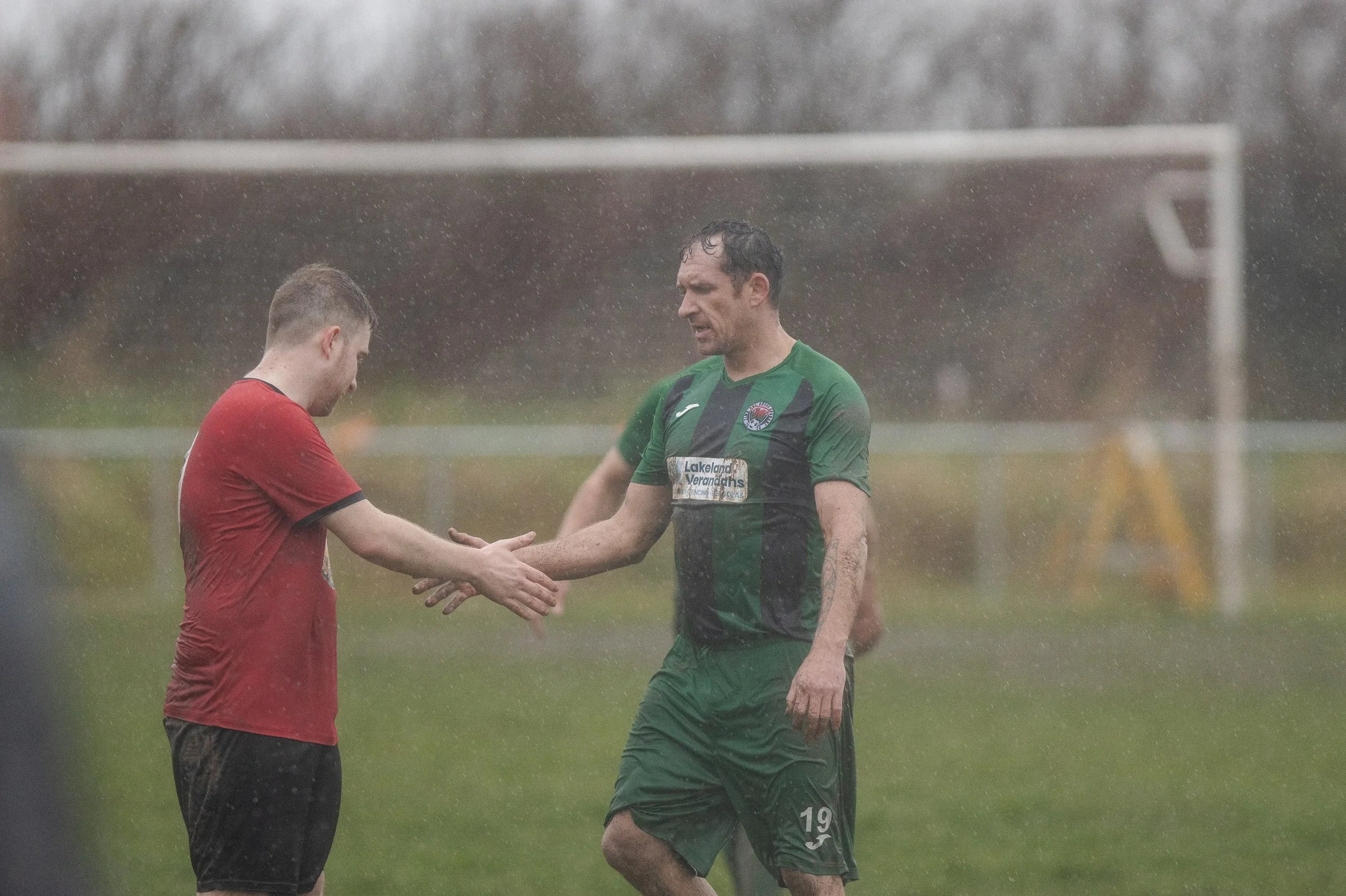 Two soccer players shake hands on a rainy field, one in a red shirt and black shorts, the other in a green and black uniform, with a soccer goal in the background.
