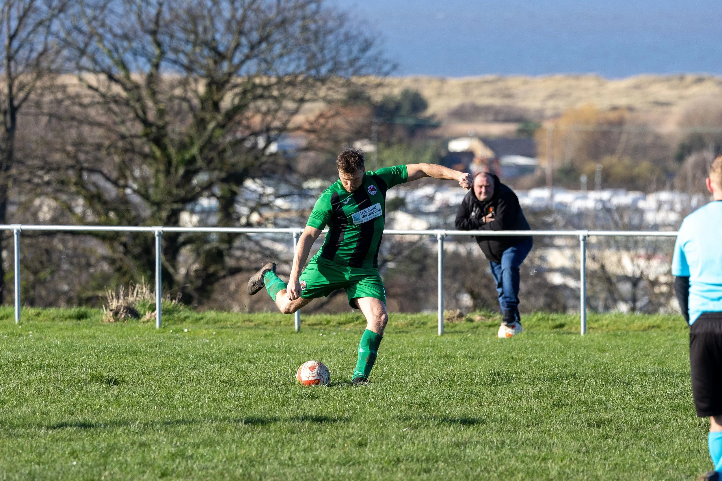A soccer player in green uniform kicking a ball during a match on a grassy field, with a man watching behind a fence and trees in the background.
