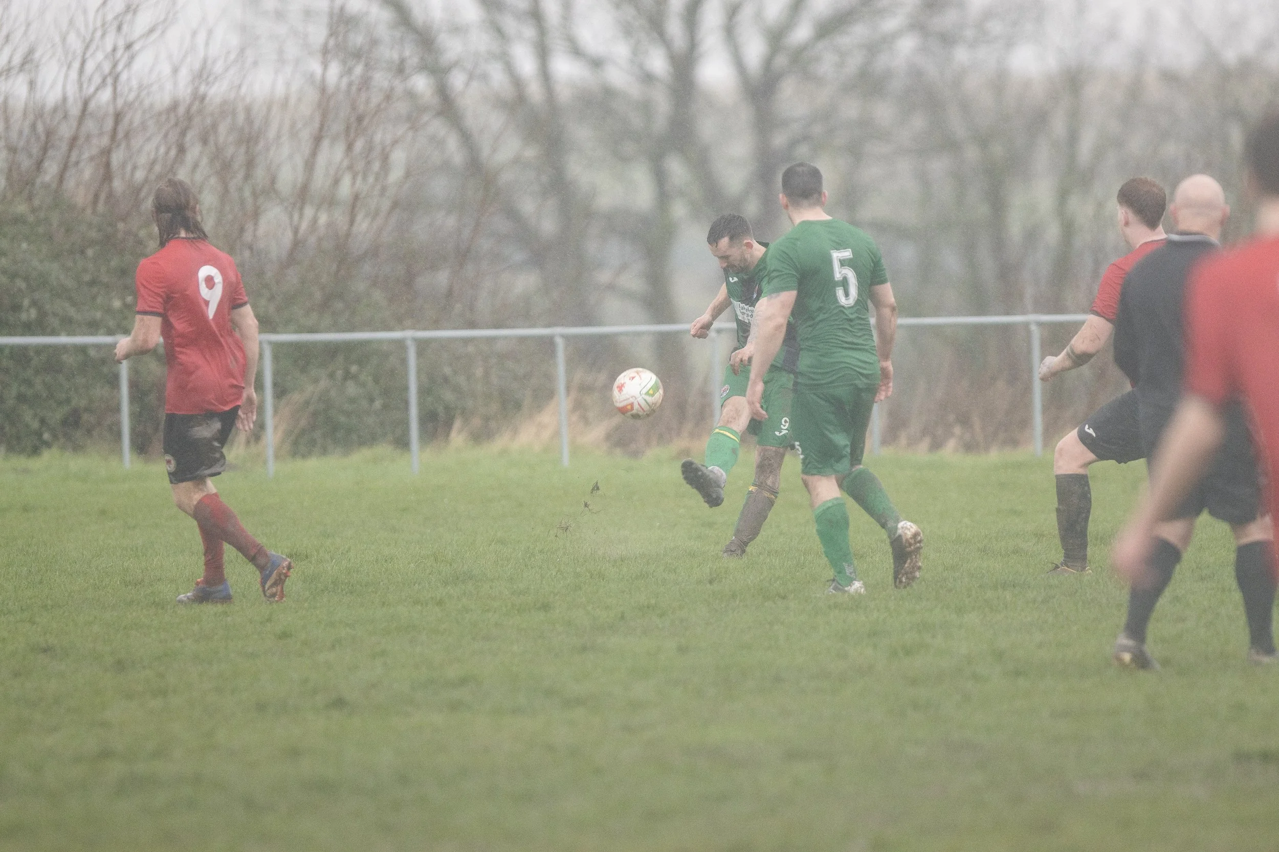Soccer match with players in red and green jerseys on a grassy field, some players actively chasing the ball.
