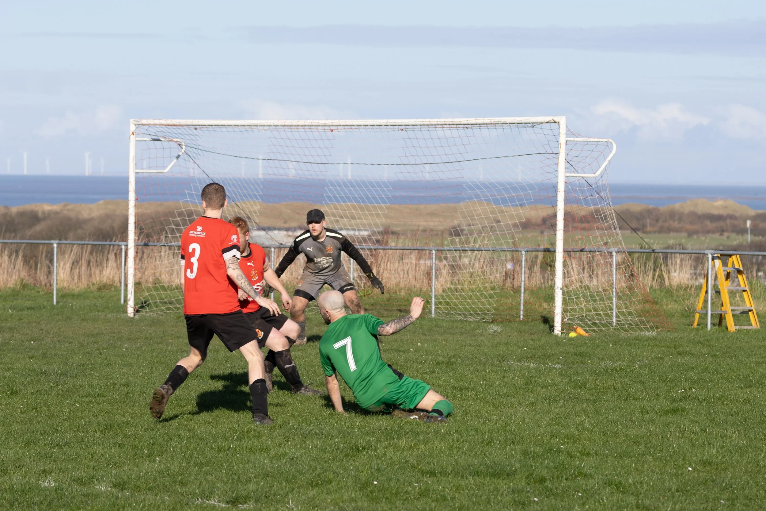Soccer players contesting the ball near the goal, with a goalkeeper preparing to block a shot on a grassy field with a coastal landscape in the background.