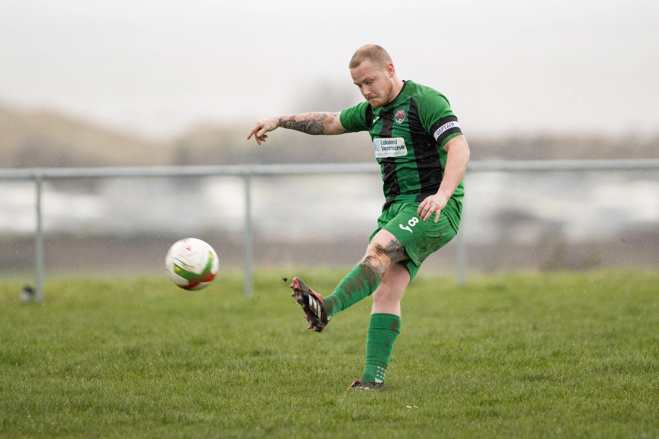A soccer player in a green and black uniform kicking a soccer ball on a grassy field during a rainy day.
