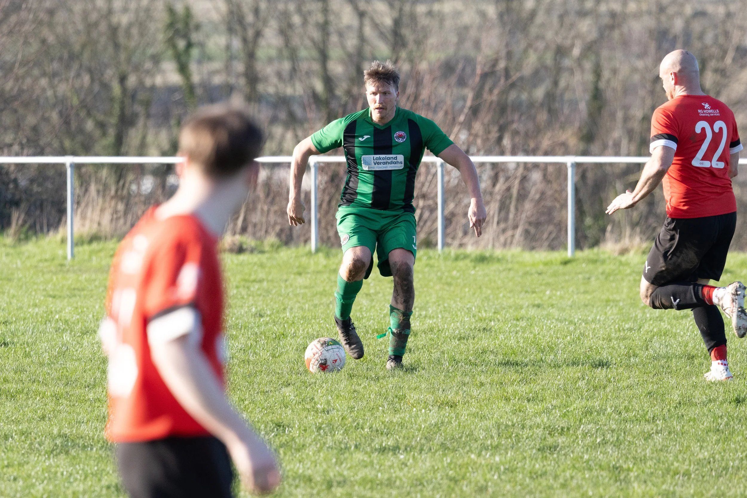 A soccer match with players on a grassy field, one in a green uniform controlling the ball while others in red uniforms are nearby.