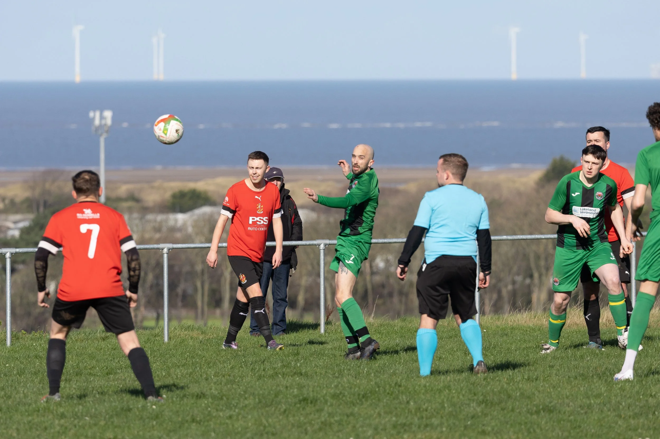 Soccer players in red and green uniforms playing a game on a grass field, with a blue sky, water, and wind turbines in the background.