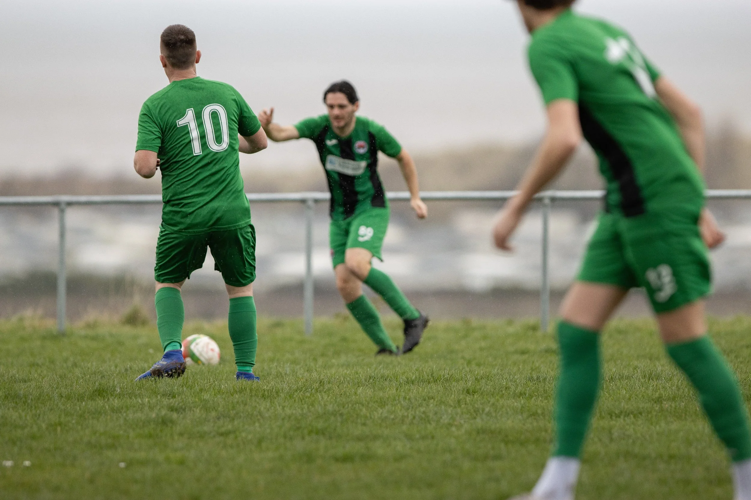 Soccer players in green uniforms playing on a grassy field, with one player preparing to kick the ball and others approaching, on a cloudy day.
