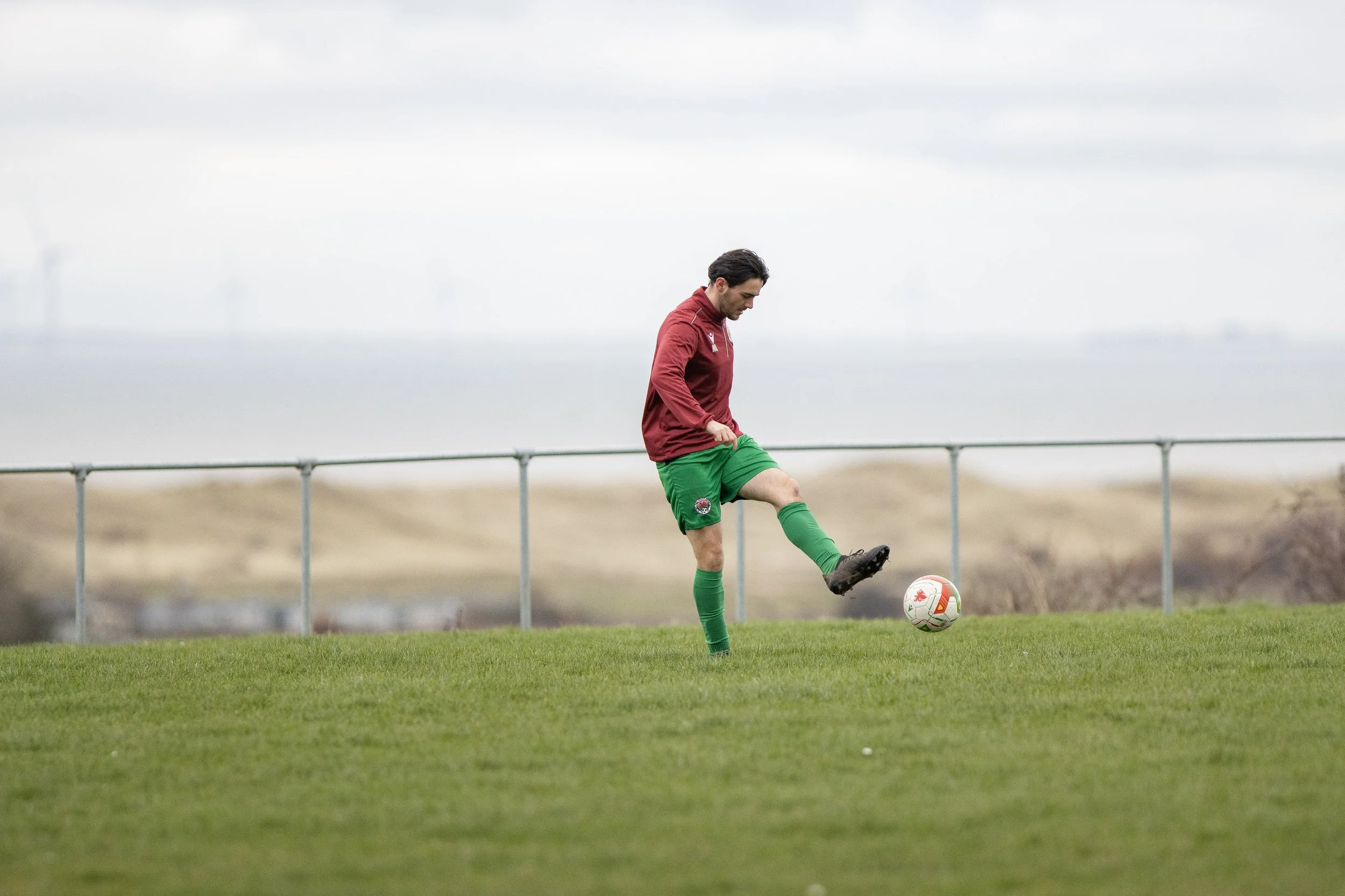 A male soccer player in a red jersey, green shorts, and green socks is on a field, controlling a soccer ball with his right foot. The background shows a cloudy sky and a distant landscape with wind turbines.