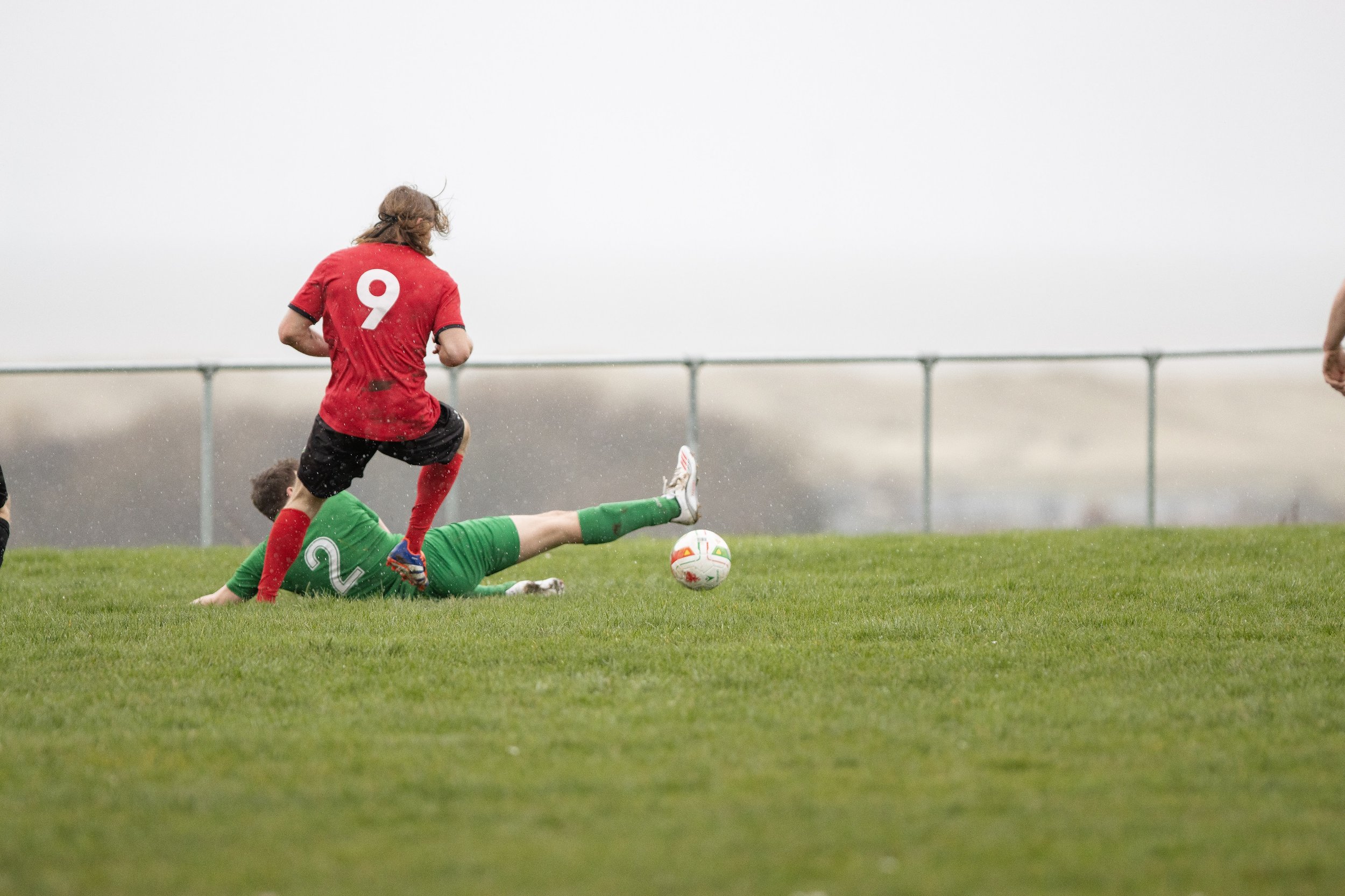 A soccer game with a player in a red jersey number 9 running while a goalkeeper in a green uniform is making a sliding save on a wet grass field during rainy weather.