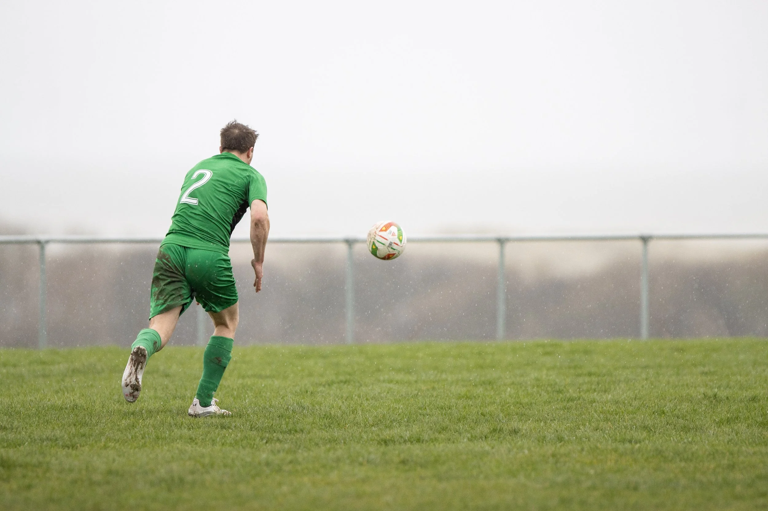 A soccer player in a green uniform with the number 2 on the back, running on a grassy field and preparing to kick a soccer ball during a rainy day.