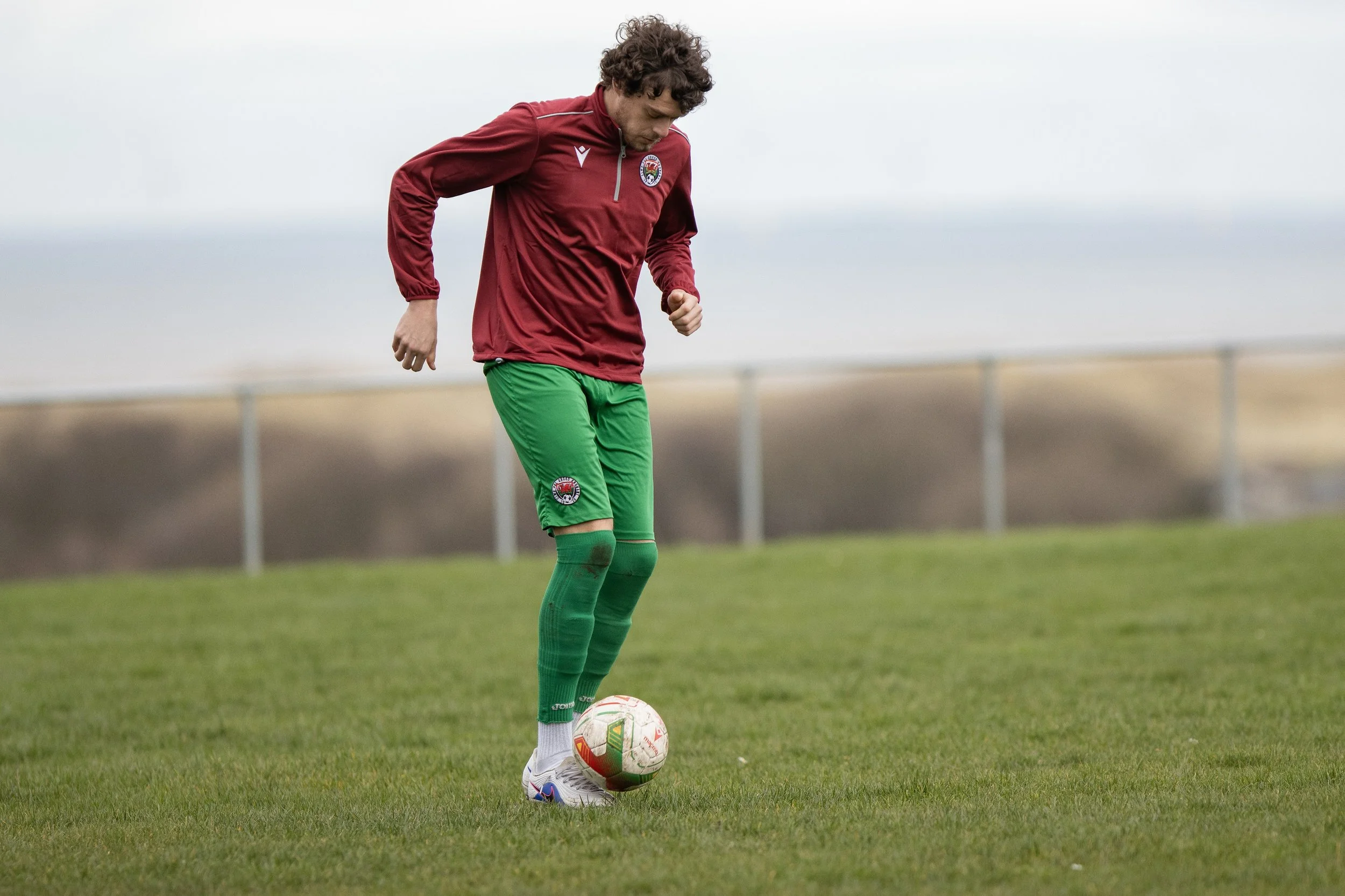 A male soccer player wearing a maroon jacket and green shorts standing on a grass field with a soccer ball