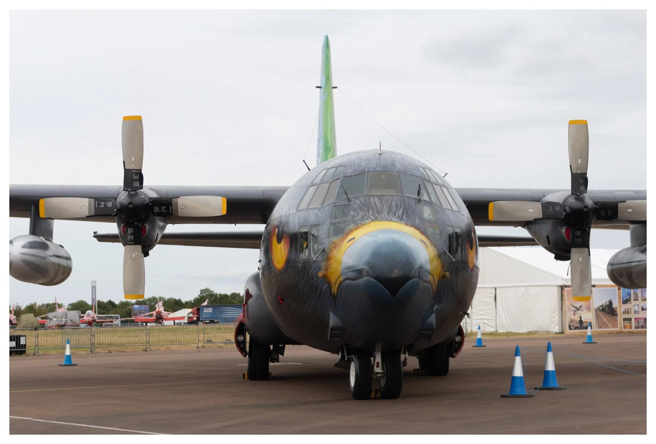 Front view of a military aircraft with a black cat design on the nose, displayed at an outdoor air show with tents and other aircraft in the background.