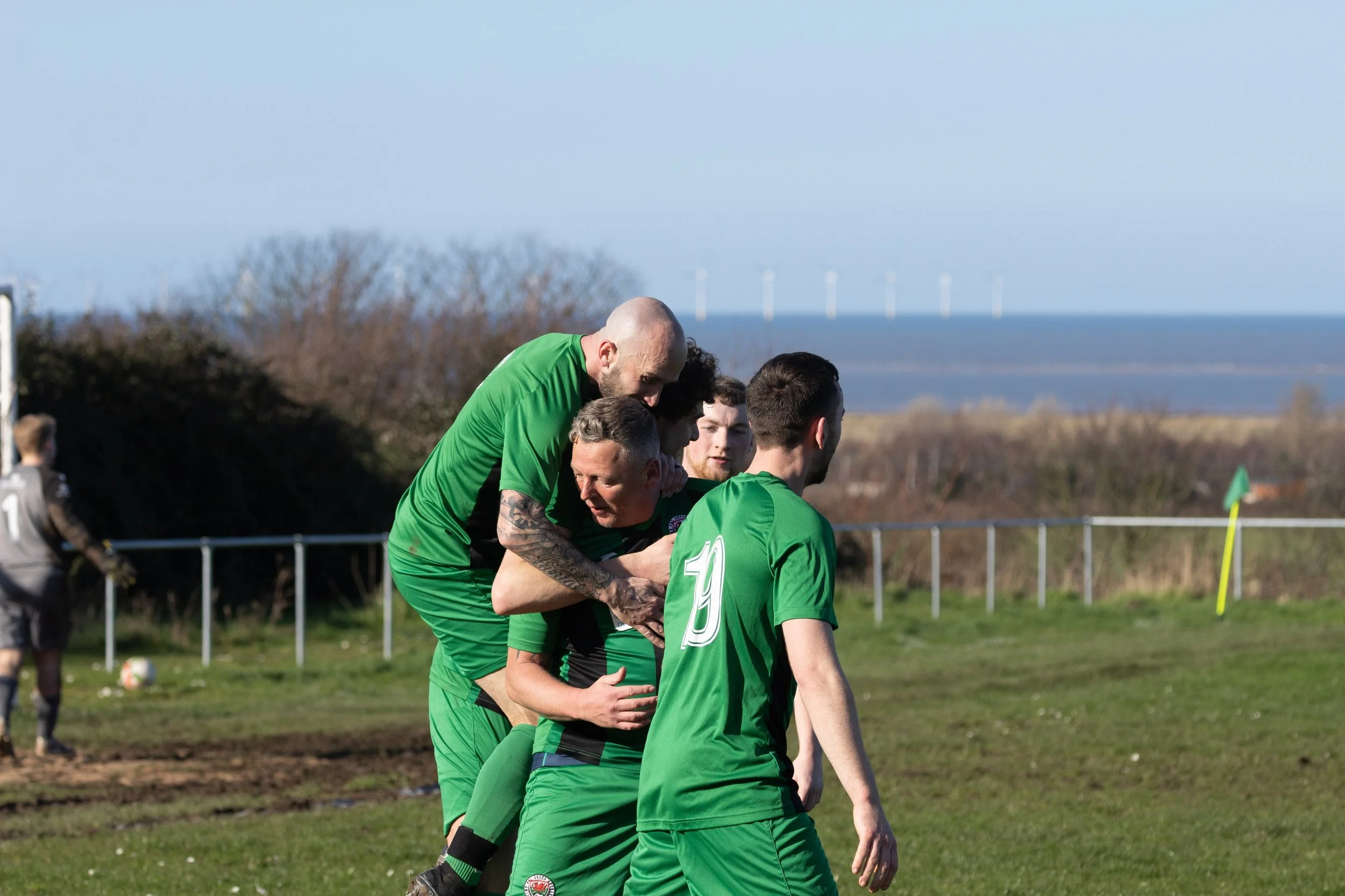 Soccer players in green jerseys celebrate a goal during a match on a grassy field with a fence, trees, and wind turbines in the background.