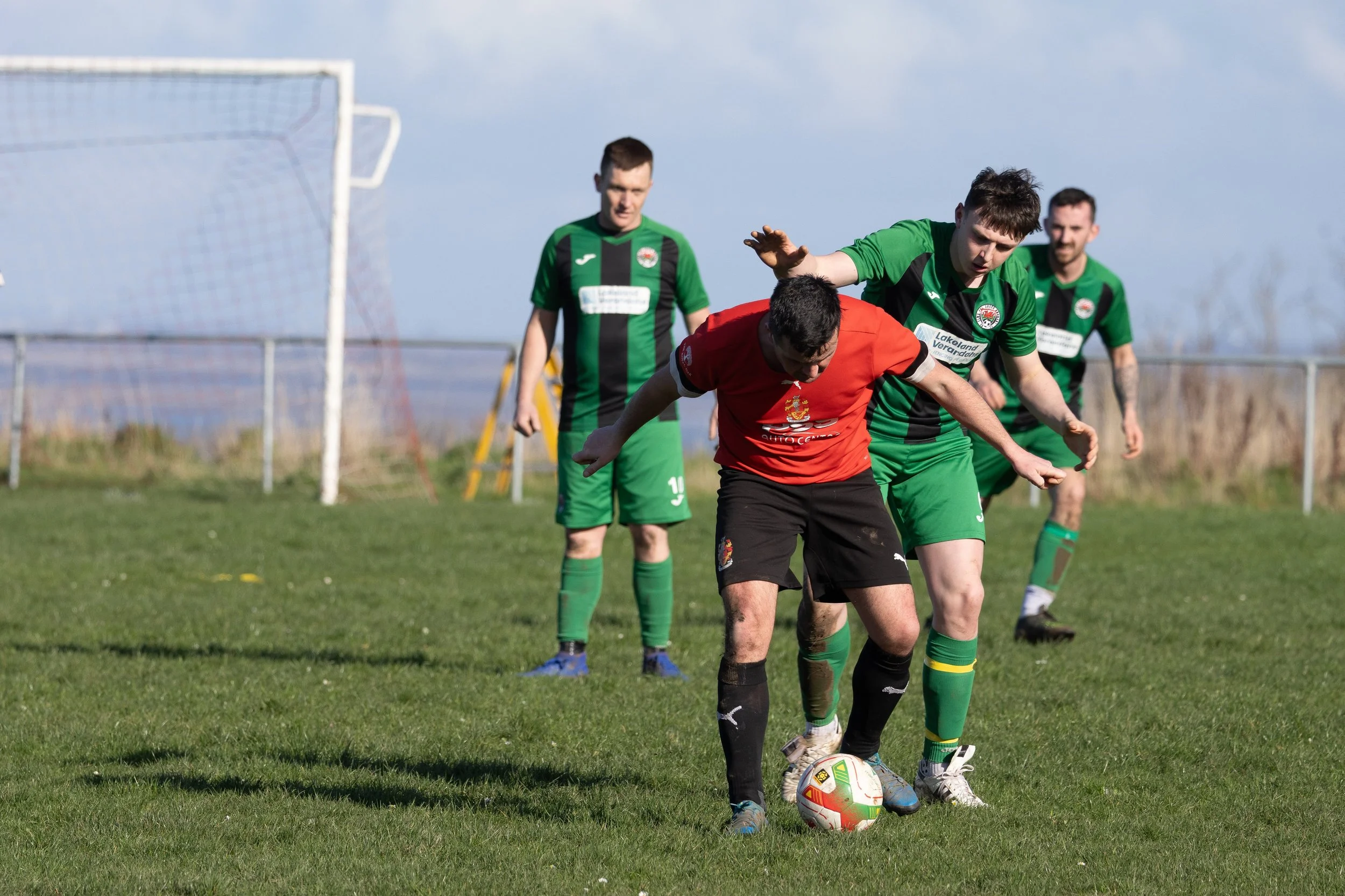 Four soccer players on a grassy field, three in green and one in red, with two players contesting the ball and one observing in the background. The players are near a goalpost, under a partly cloudy sky.