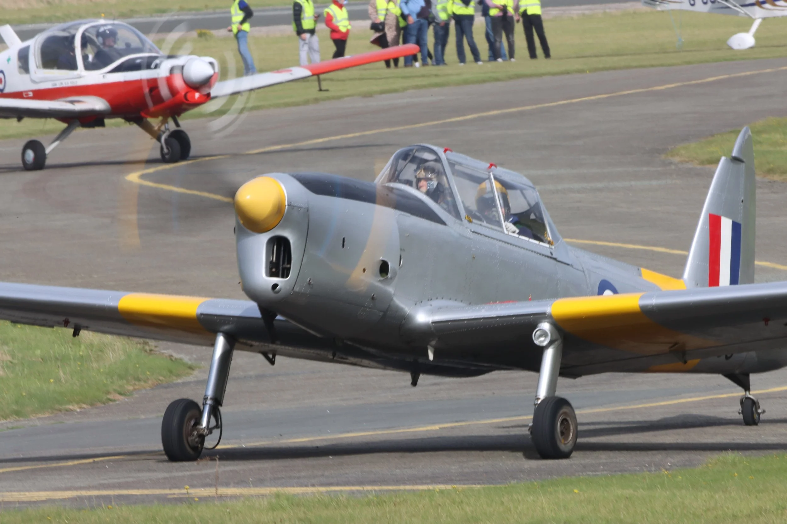 Vintage military aircraft taxiing on runway with propeller spinning, behind a second red and white aircraft, and spectators watching from the grassy area.