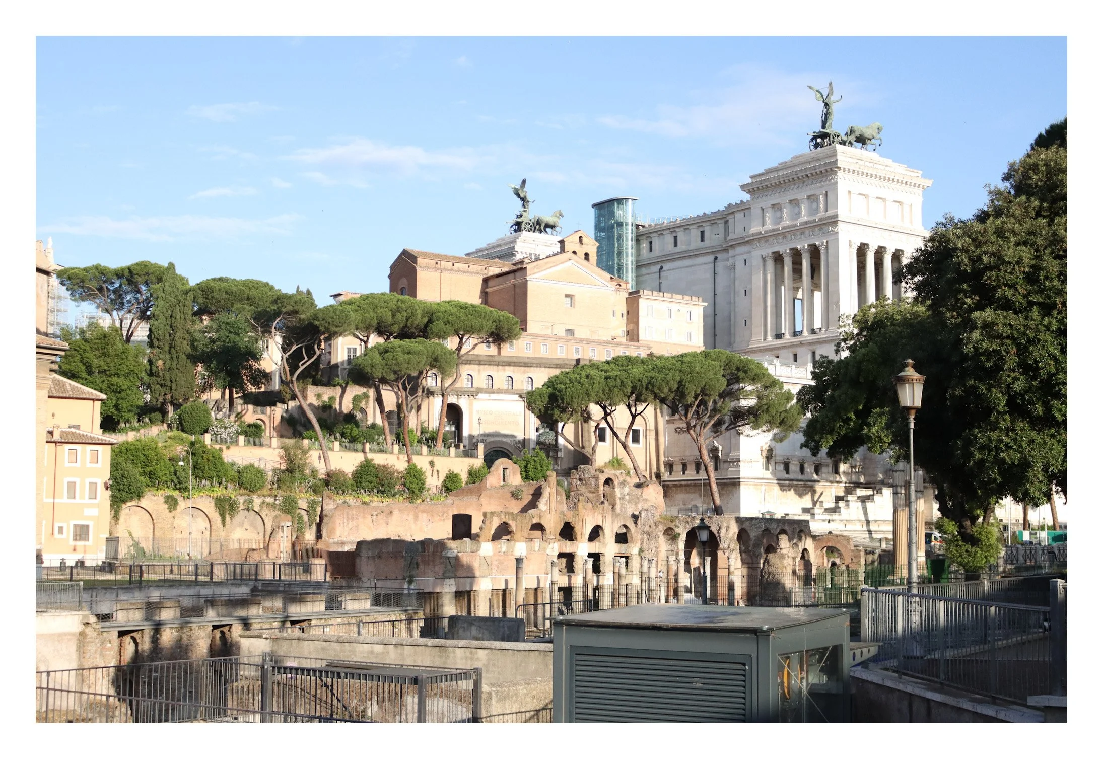 A cityscape of Rome, Italy, featuring ancient ruins in the foreground, lush green trees, and historical buildings including the Vittorio Emanuele II Monument with statues on top, under a clear blue sky.