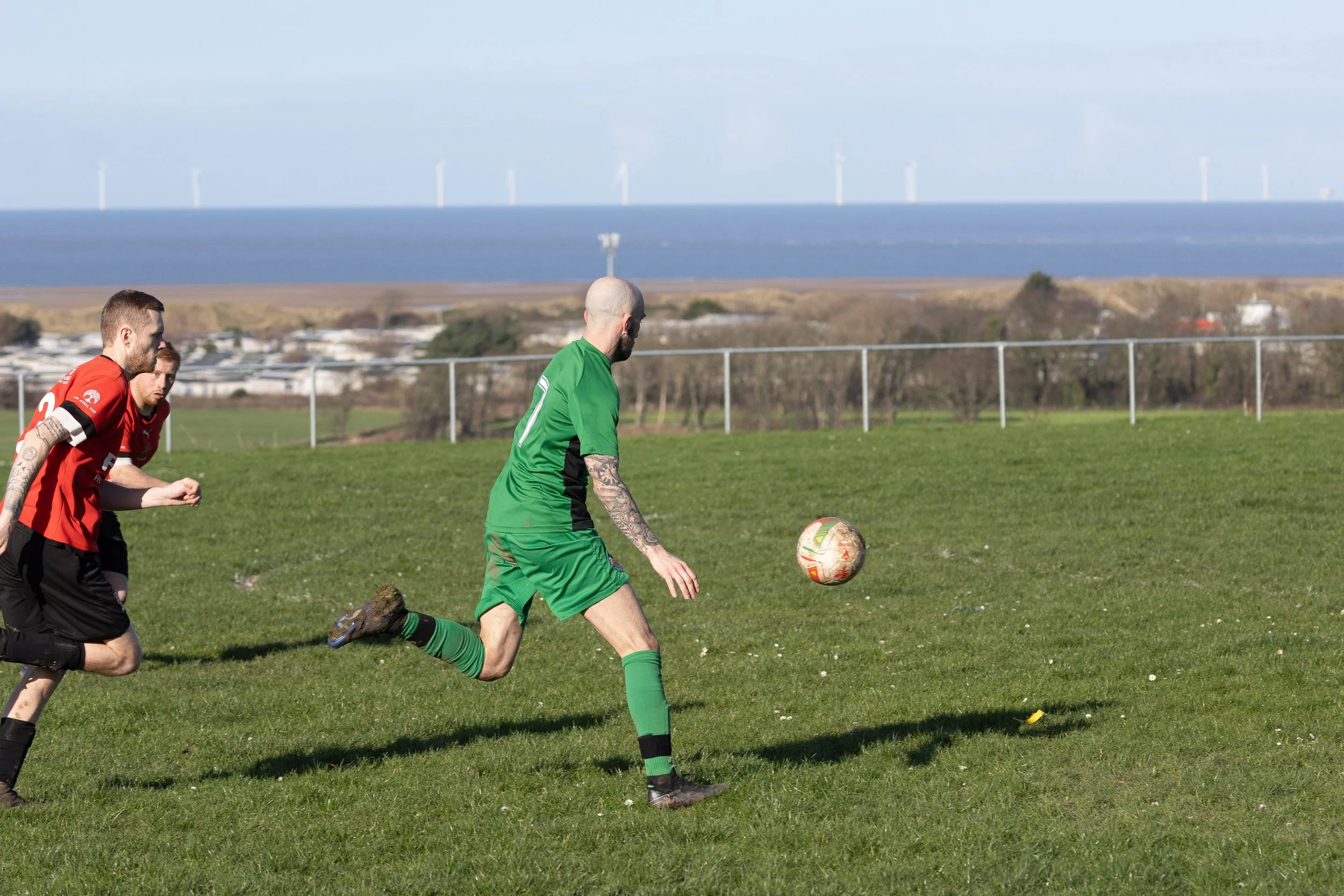 Two soccer players chasing a ball on a field with a fence and ocean in the background.