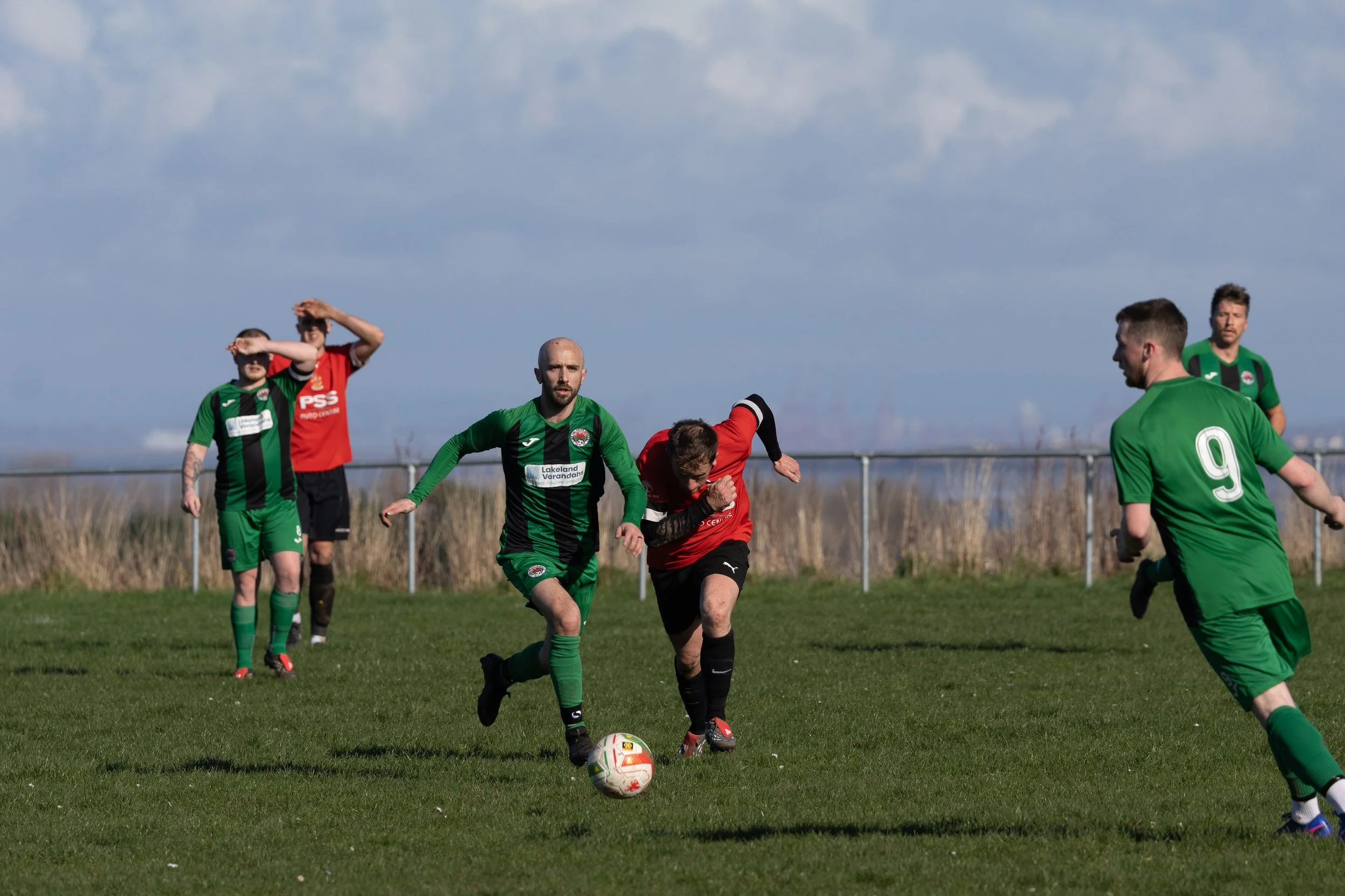 Soccer match with players in green and red jerseys on a grassy field under a cloudy sky.