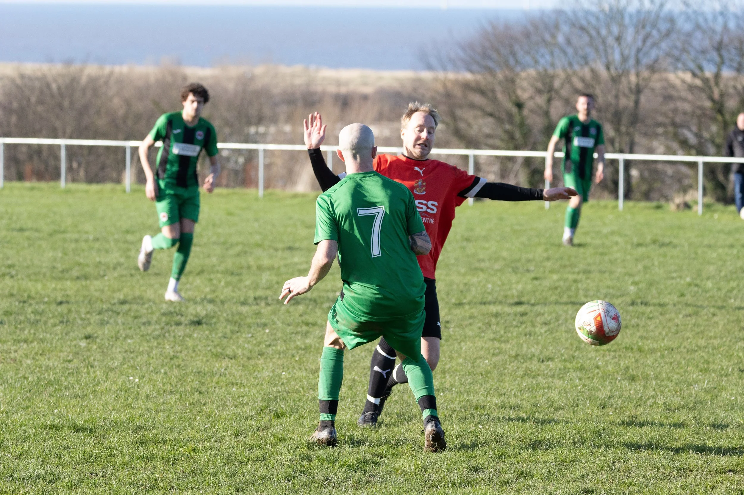 Soccer players in action on a grassy field, with two players near the foreground contesting for the ball, one in green and the other in red, and three players in the background watching play.