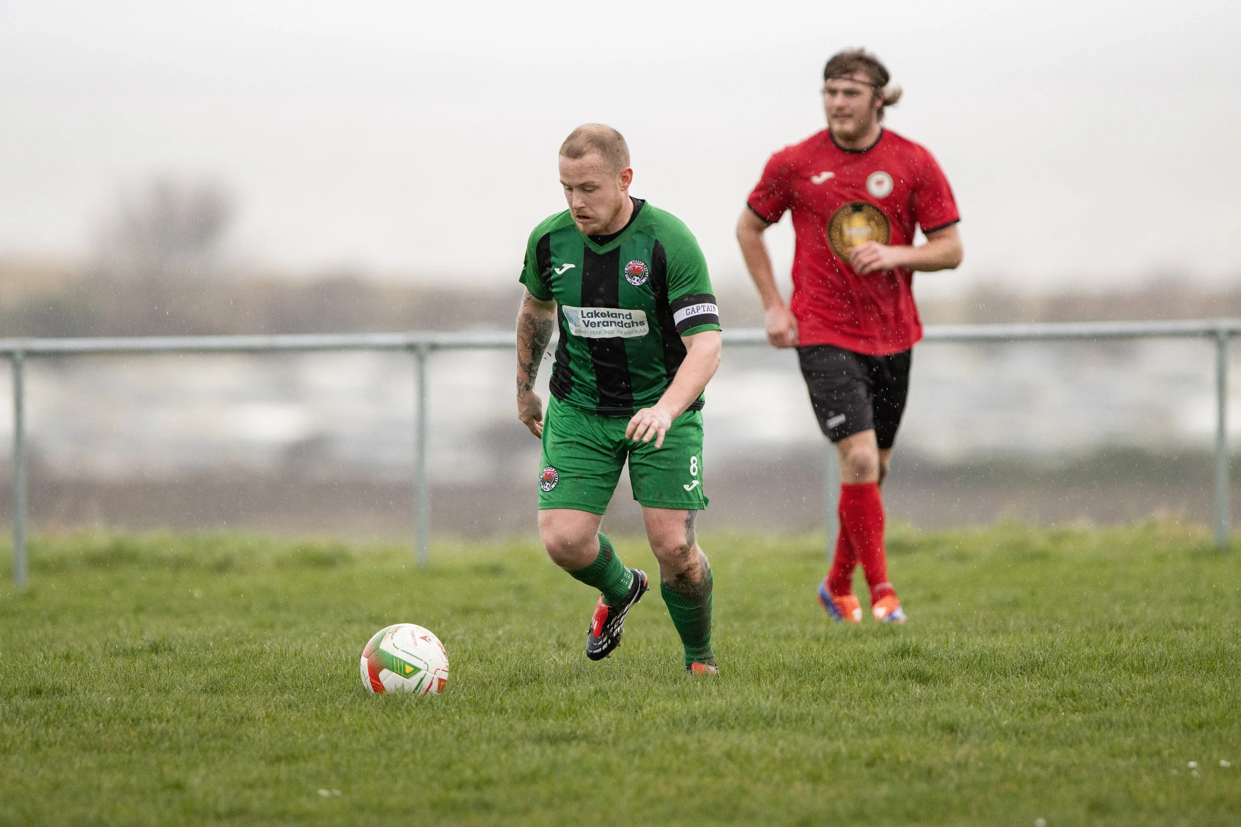 A soccer player in a green and black uniform is kicking a soccer ball on a grassy field, with another player in a red uniform running in the background under rainy weather.