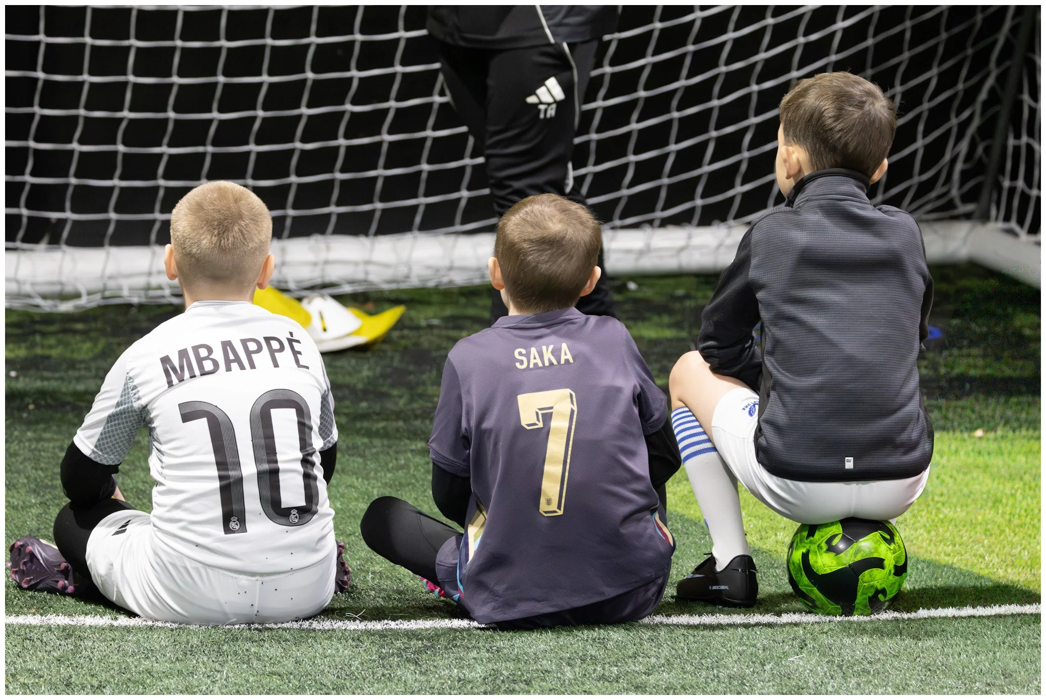 Three young boys sitting on a soccer field, watching a person in black athletic clothing near a goal net. The boy on the left wears a white jersey with 'MBAPPÉ' and the number 10, the middle boy wears a dark jersey with 'SAKA' and the number 7, and t