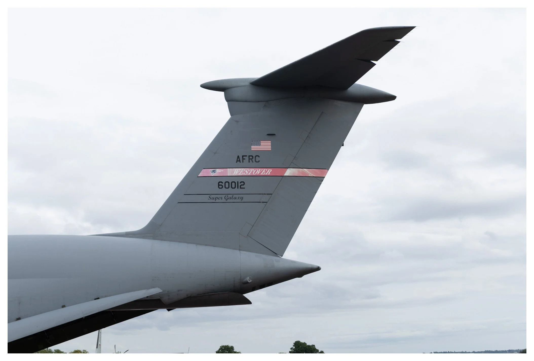 Close-up of the tail section of a military aircraft featuring the US flag, identification markings, and a pink strip labeled "Westover," with a cloudy sky in the background.