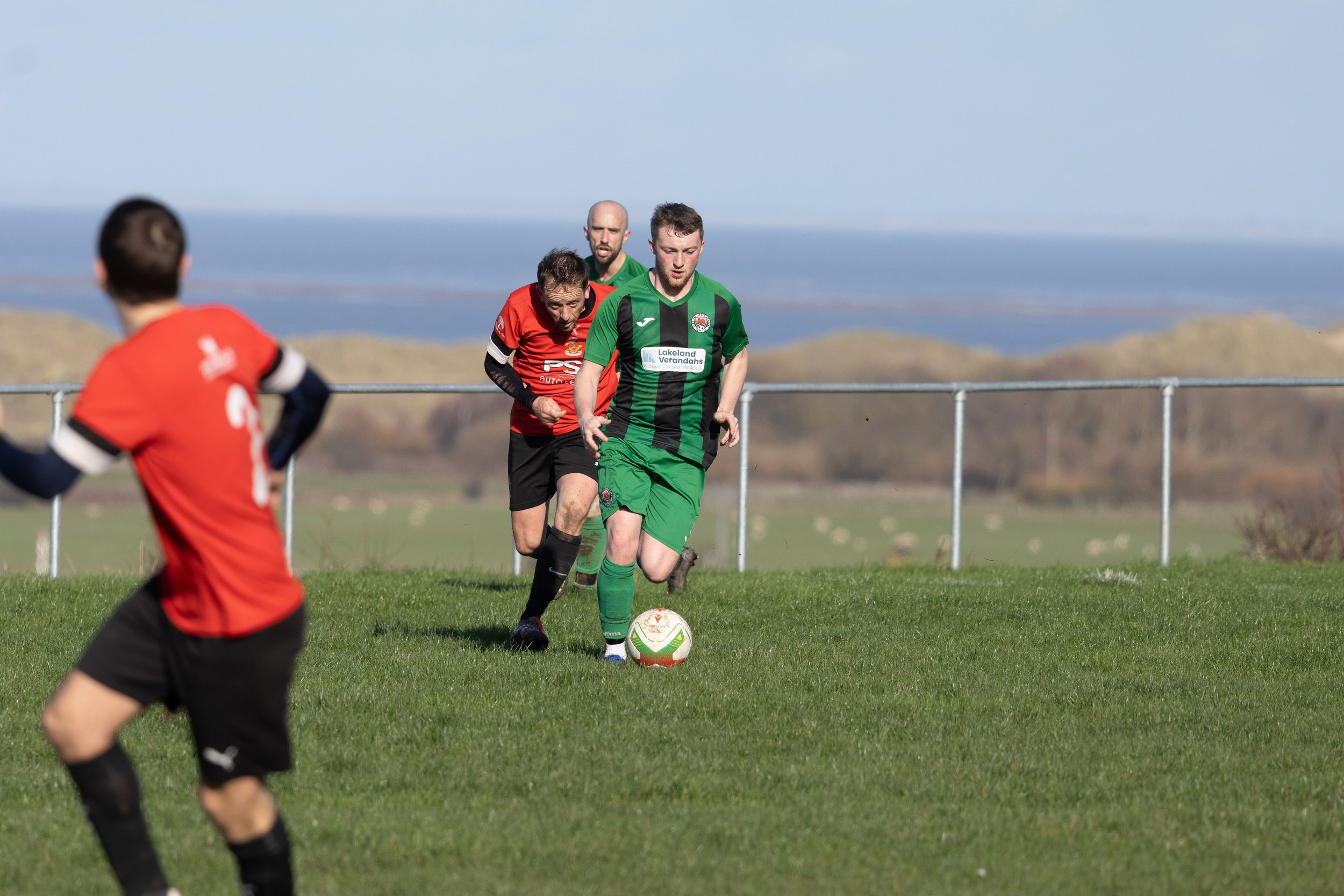 Soccer players in action on a grassy field with a scenic background of hills and sky, including a player in green controlling the ball and others in red nearby.