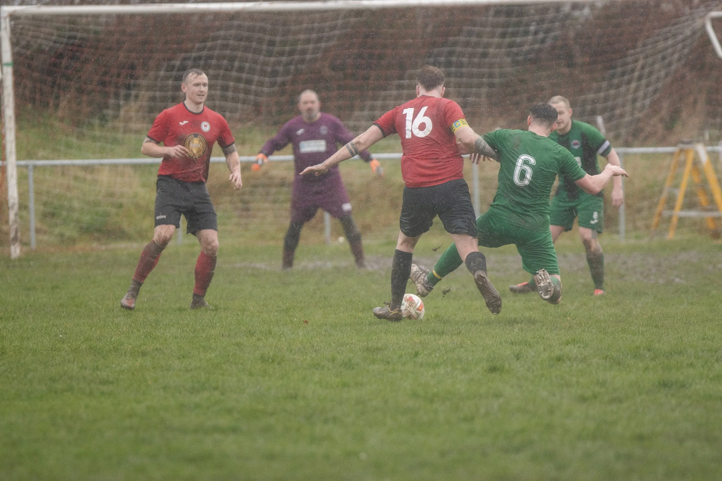 Soccer game in rainy weather with players competing for the ball, one player in a green jersey being tackled by a player in a red jersey, three other players and a goalkeeper watching in the background.