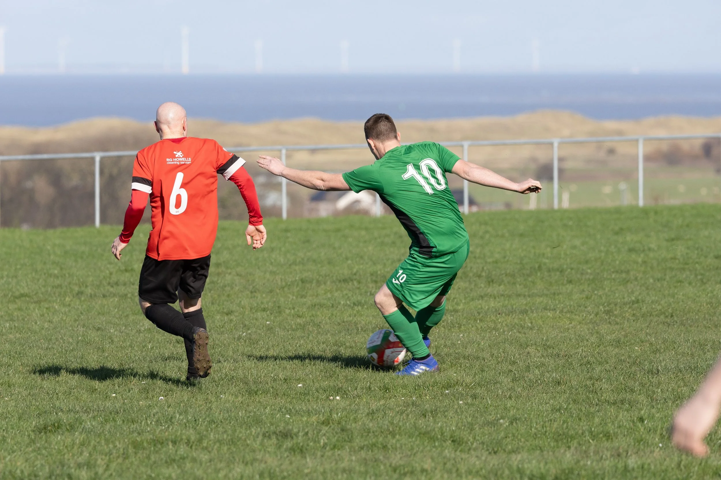 Two soccer players on a grassy field, one in a red jersey with the number 6, the other in a green jersey with the number 10, competing for the ball. The player in green is reaching out with his right arm, while the player in red is approaching from b