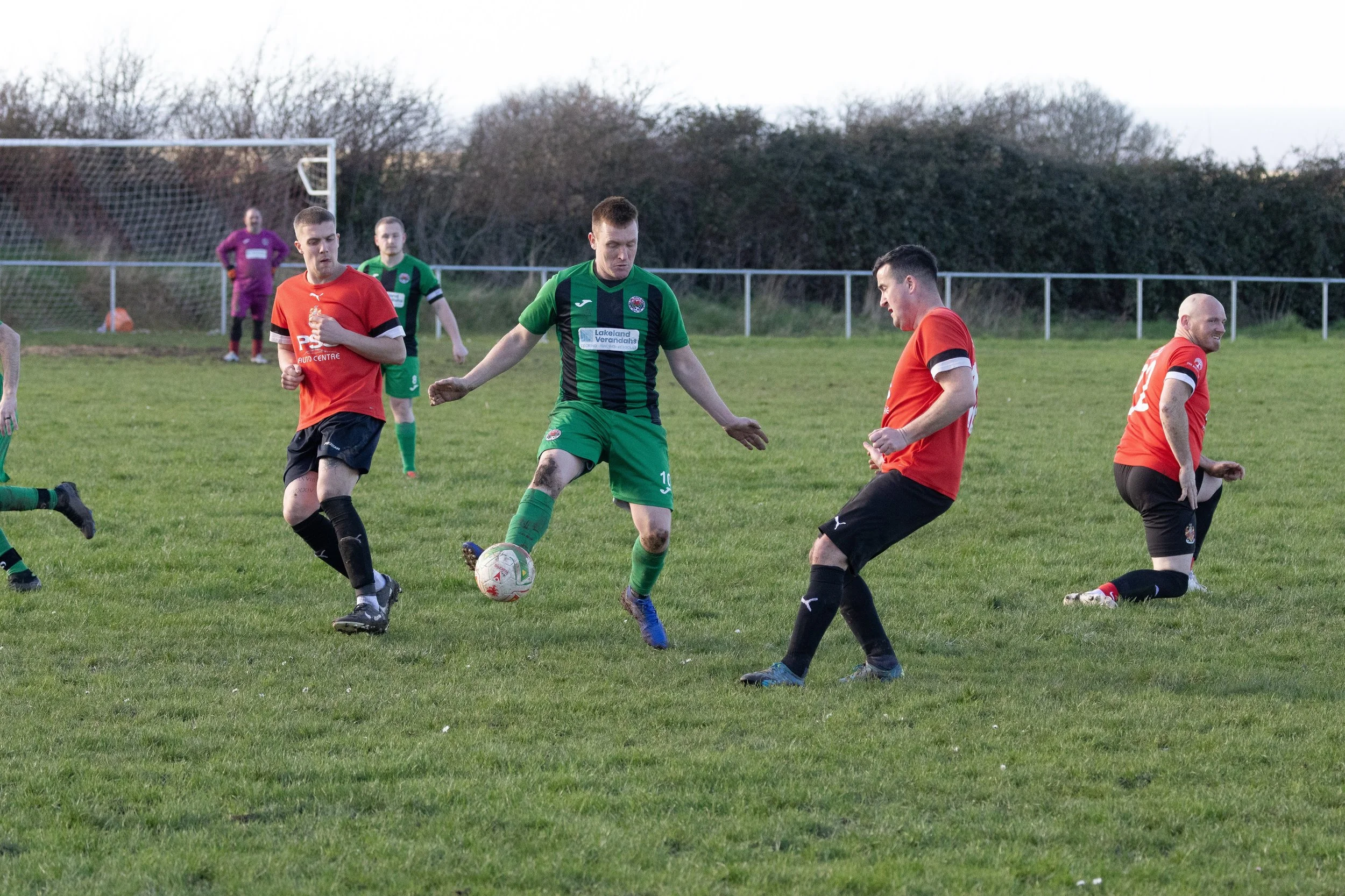 Soccer players on a field during a match, with a player in a green and black uniform controlling the ball while others in red and black attempt to block him. Goalpost and additional players in the background.