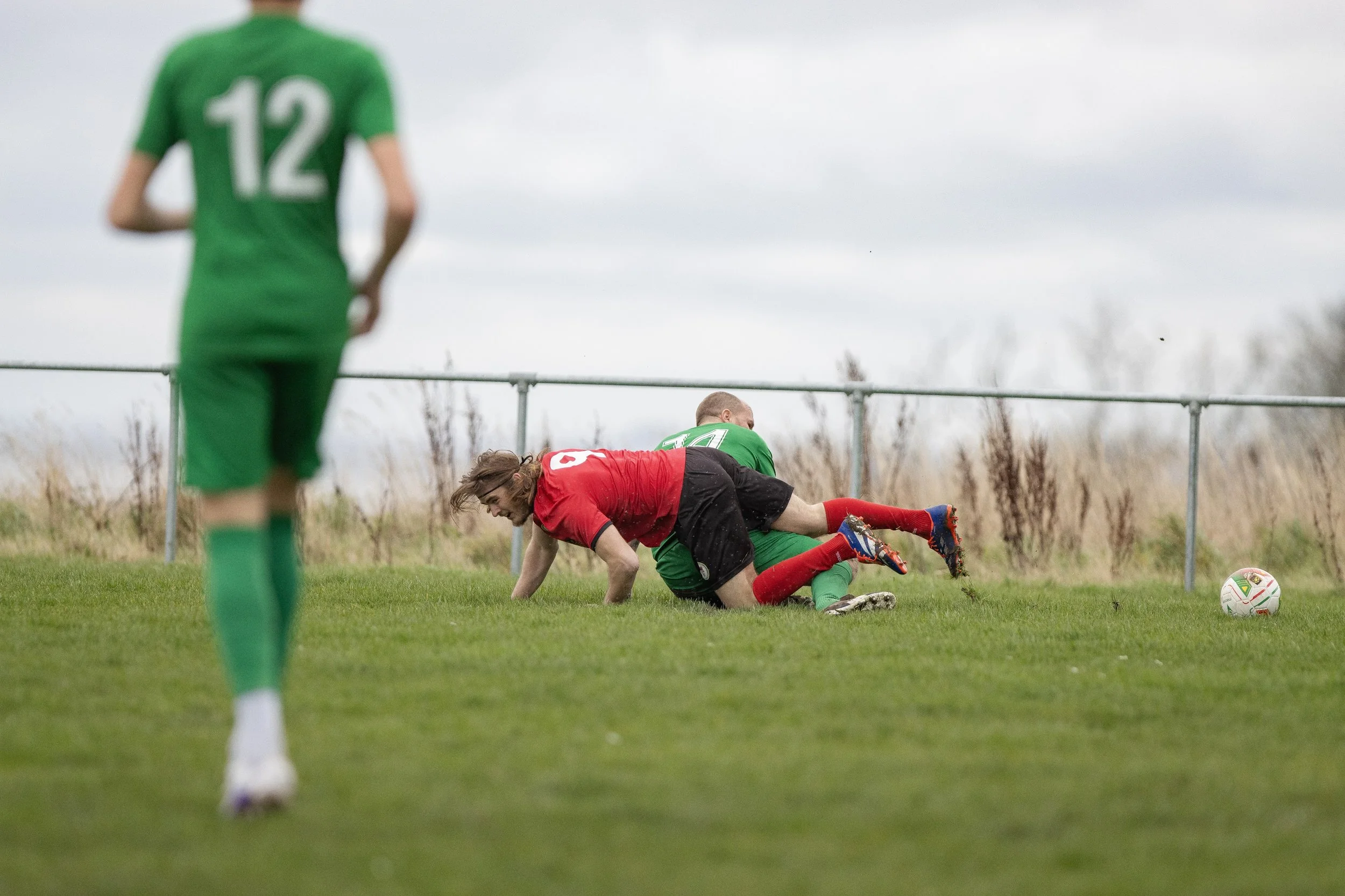Two soccer players, one in red and black and the other in green, are on the ground battling for the ball during a match, with a third player in green watching nearby on a grassy field under cloudy skies.