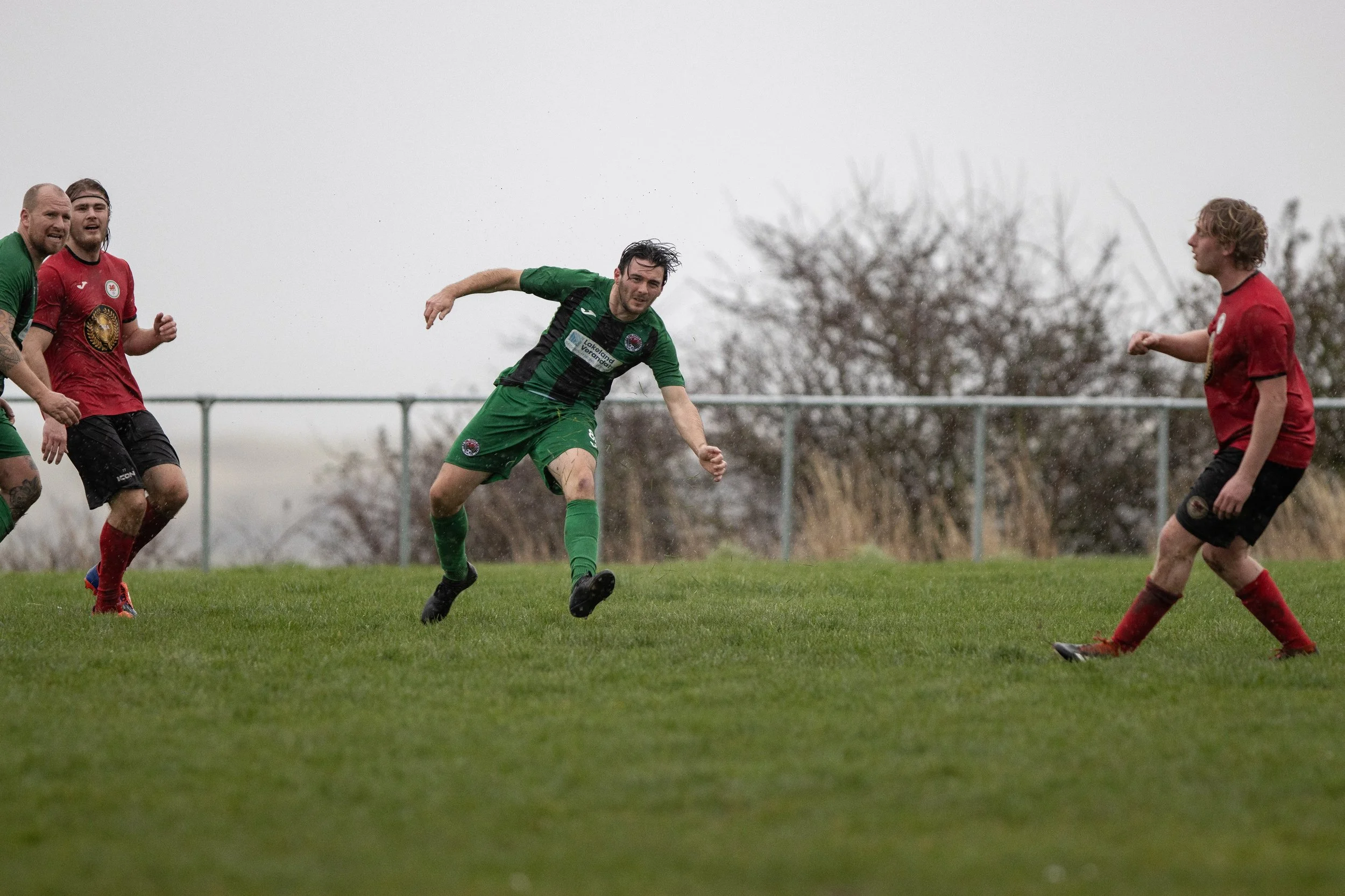 Soccer players in action on a rainy day, with a player in green running towards the right and others in red near him on a grassy field.