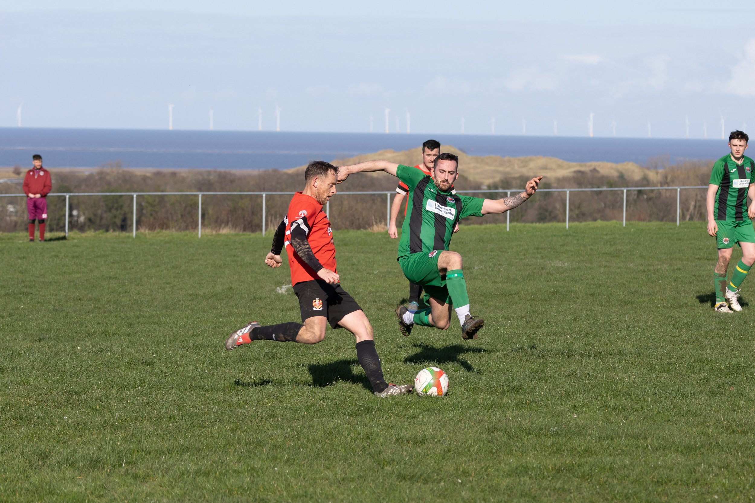 Soccer players competing for the ball on a grassy field with a coastal landscape and wind turbines in the background.