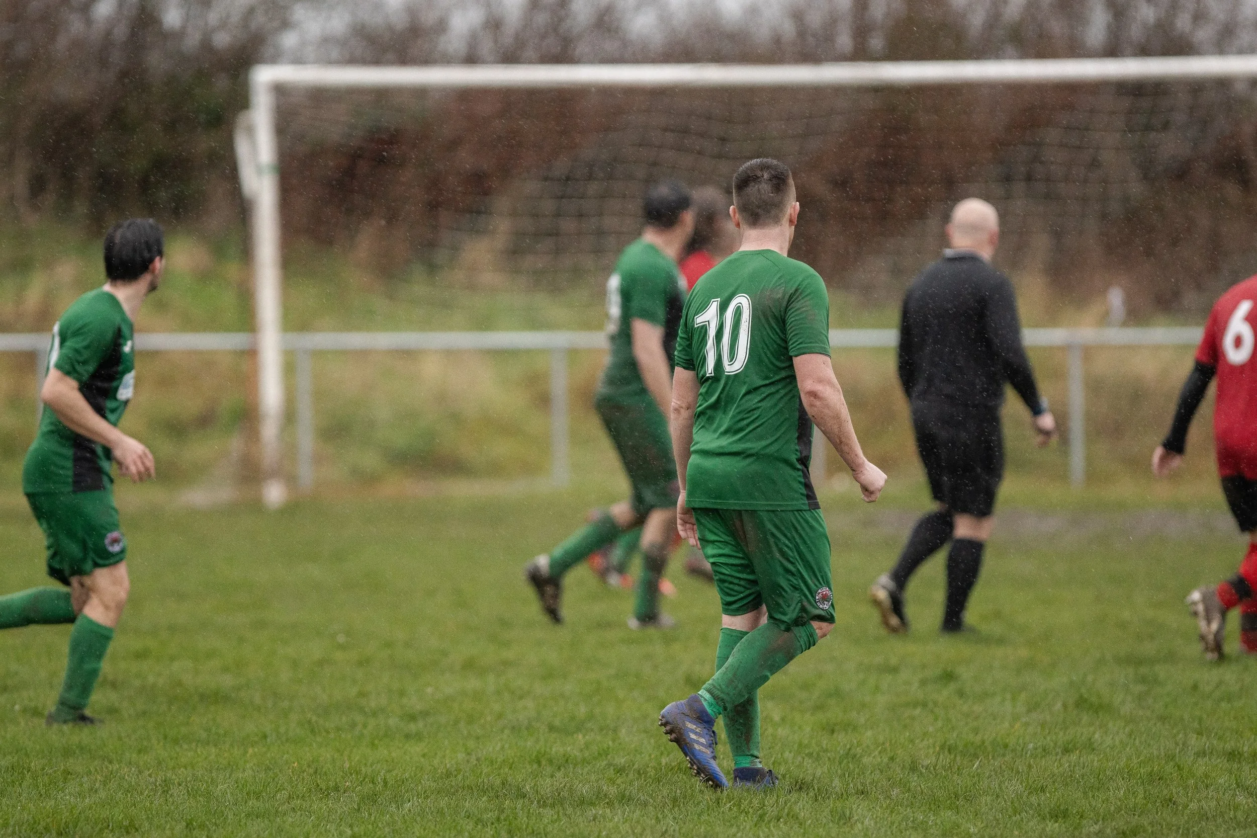 Soccer players on a rainy field, with one player wearing a green jersey with the number 10, and a few others in green and red jerseys, near the goal post.