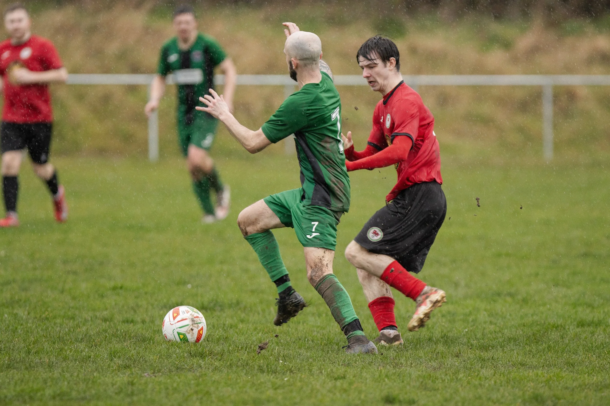 Two soccer players, one in a green uniform and the other in a red uniform, compete for the ball on a rain-soaked field. The green player is trying to control the ball while the red player attempts to challenge him. In the background, other players ar