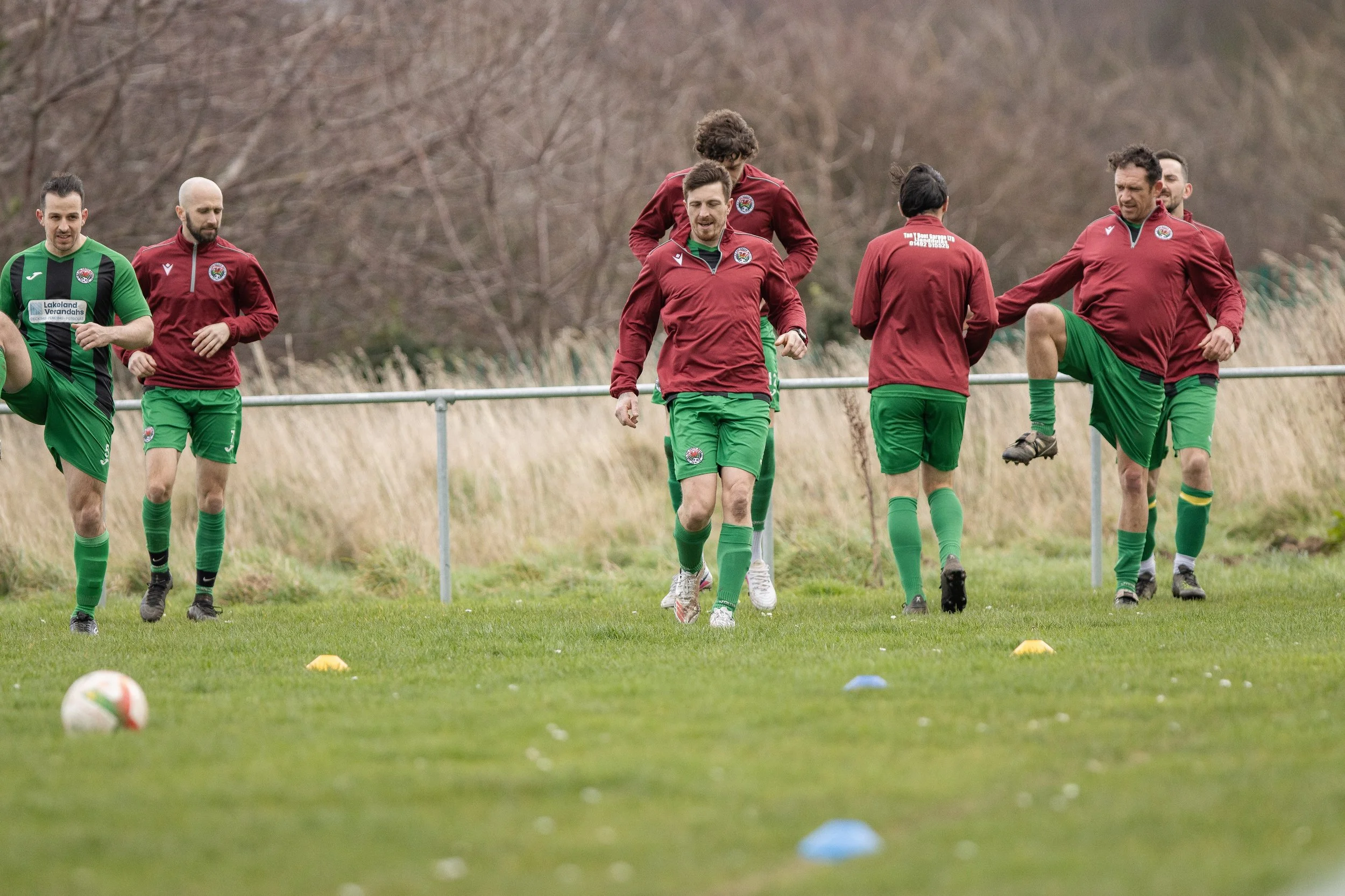 Soccer players in red and green uniforms during a training session on a grassy field, with cones set up for drills and trees in the background.