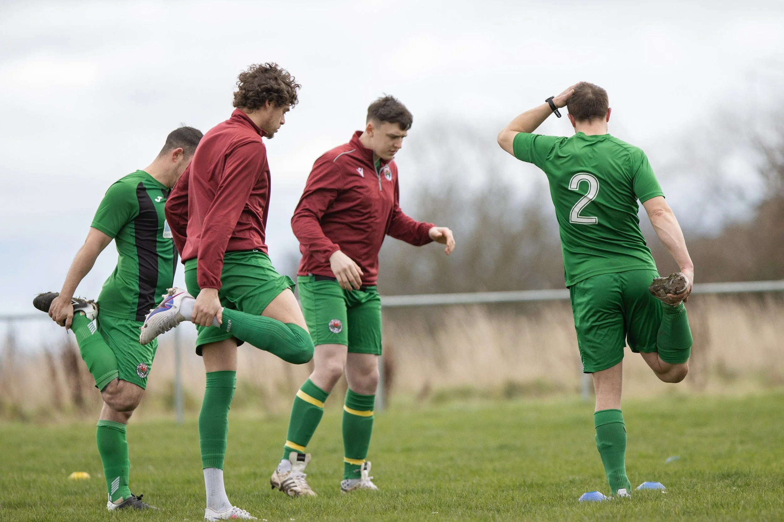 Four male soccer players stretching on a grassy field, with three wearing green shorts and one in a maroon jacket, under an overcast sky.