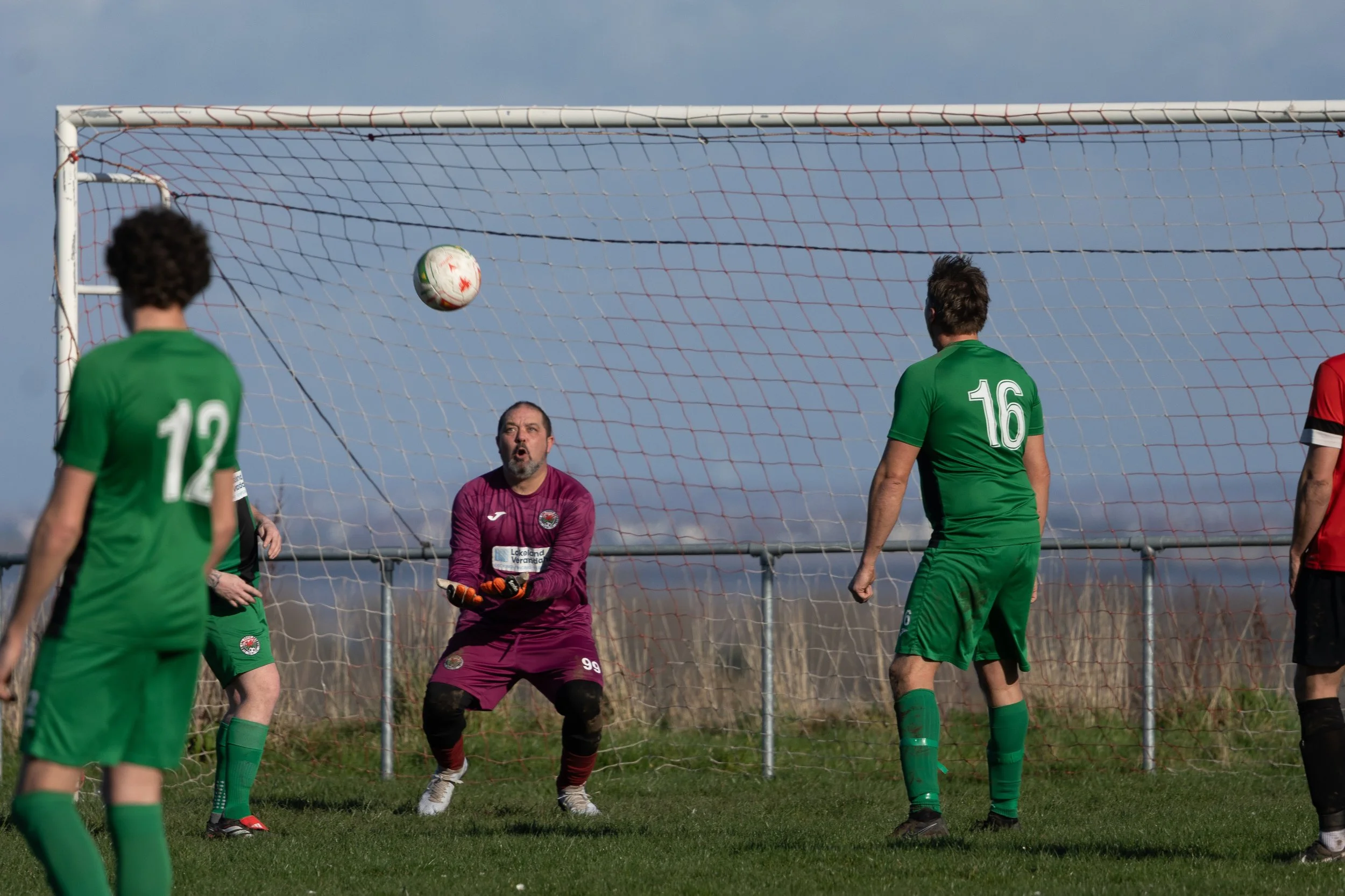 A soccer game in progress with players in green jerseys and a goalkeeper in purple, attempting to block the ball near the goal.