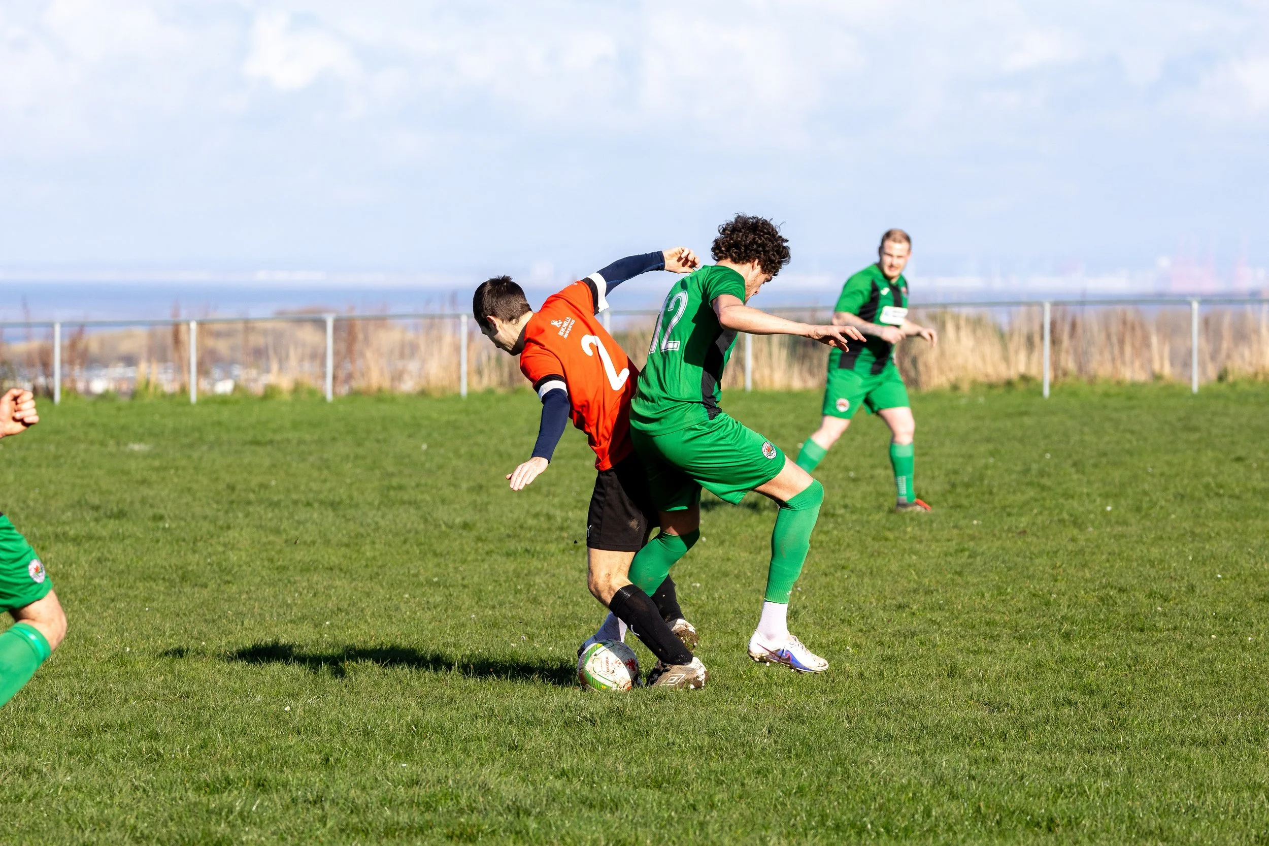 Two soccer players, one in a red jersey and the other in a green jersey, contest for the ball on a grassy field during a match, with other players and a fence in the background.