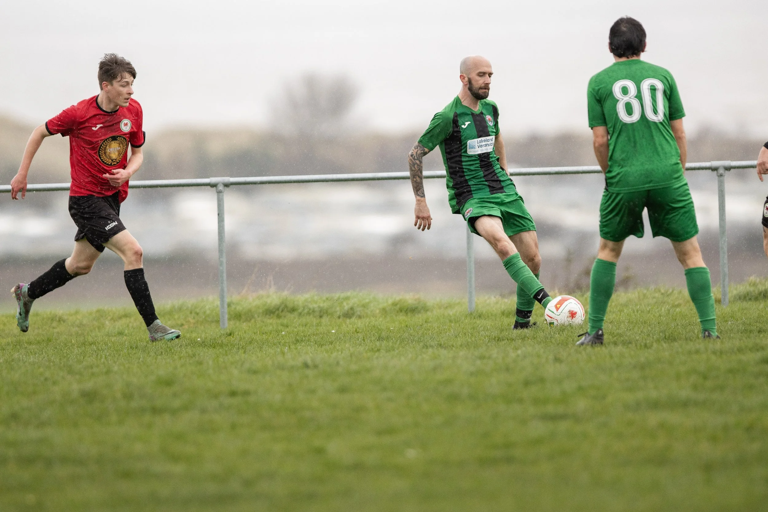 Soccer players on the field during a match, with one player in a green and black uniform controlling the ball, another player in a red jersey running, and a third in a green jersey with the number 80 standing nearby.