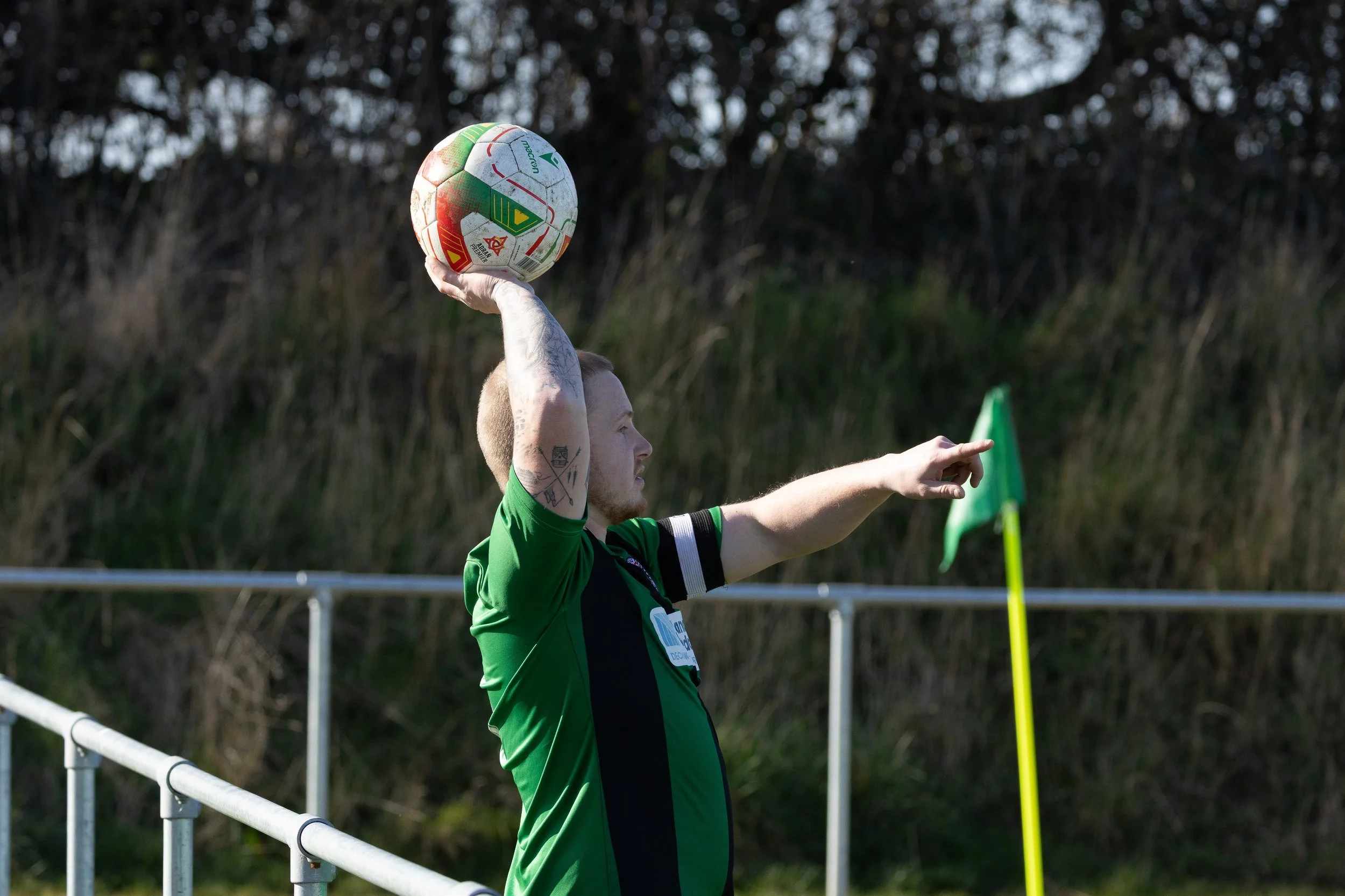 A soccer player in a green jersey holding a soccer ball overhead on the sidelines, pointing with his right arm during a game.