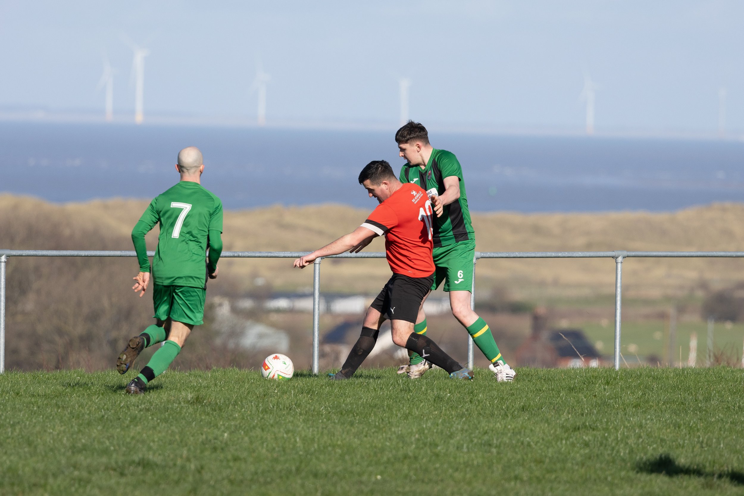 Two soccer players in green jerseys and one player in a red shirt compete for the ball on a grassy field, with wind turbines visible in the background.
