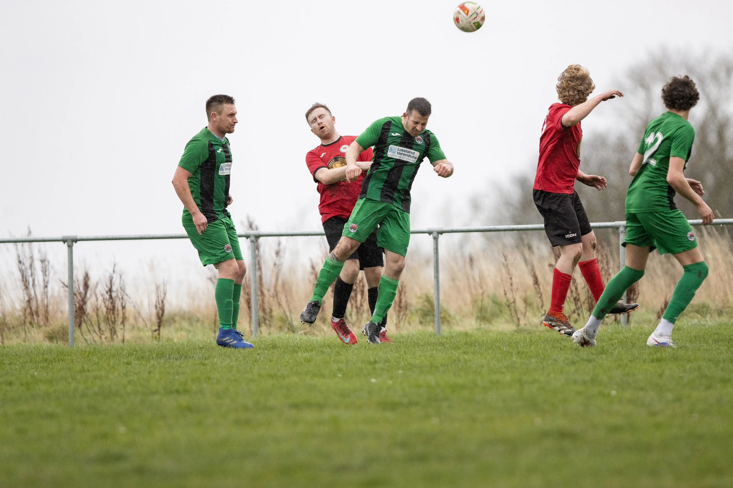 Soccer players competing for ball on field, wearing green and red uniforms, overcast day.