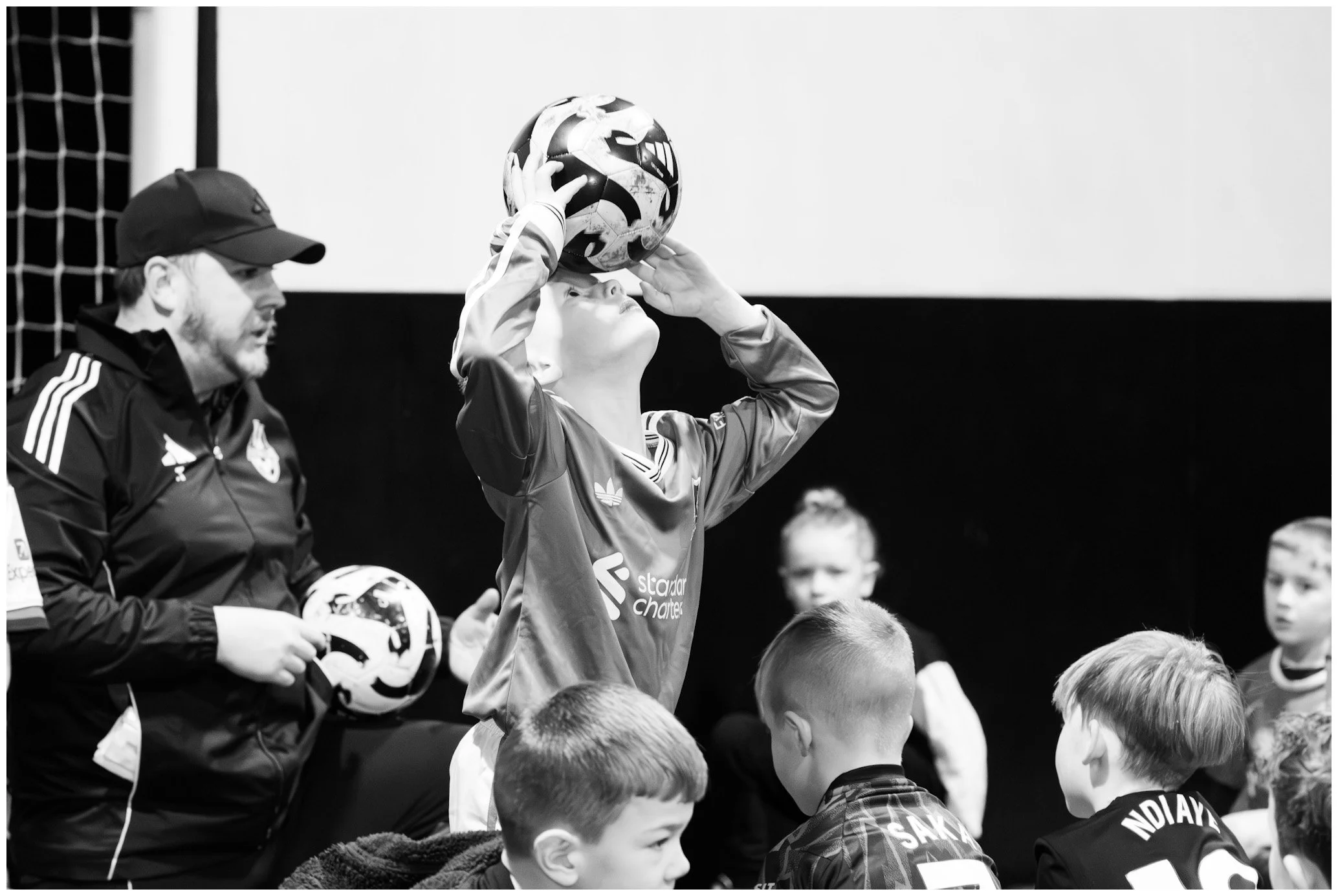 A young boy in a sports jersey holding a soccer ball above his head, surrounded by children and an adult, in an indoor sports setting.