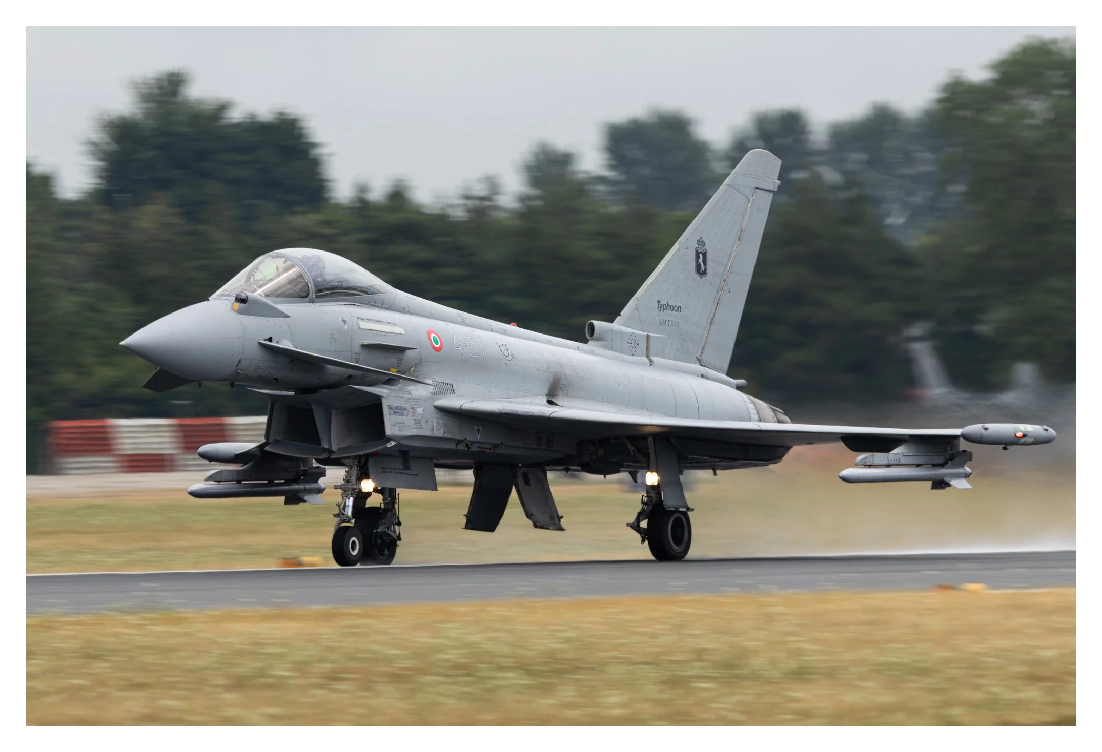 A military fighter jet aircraft taxiing on a runway with trees in the background.
