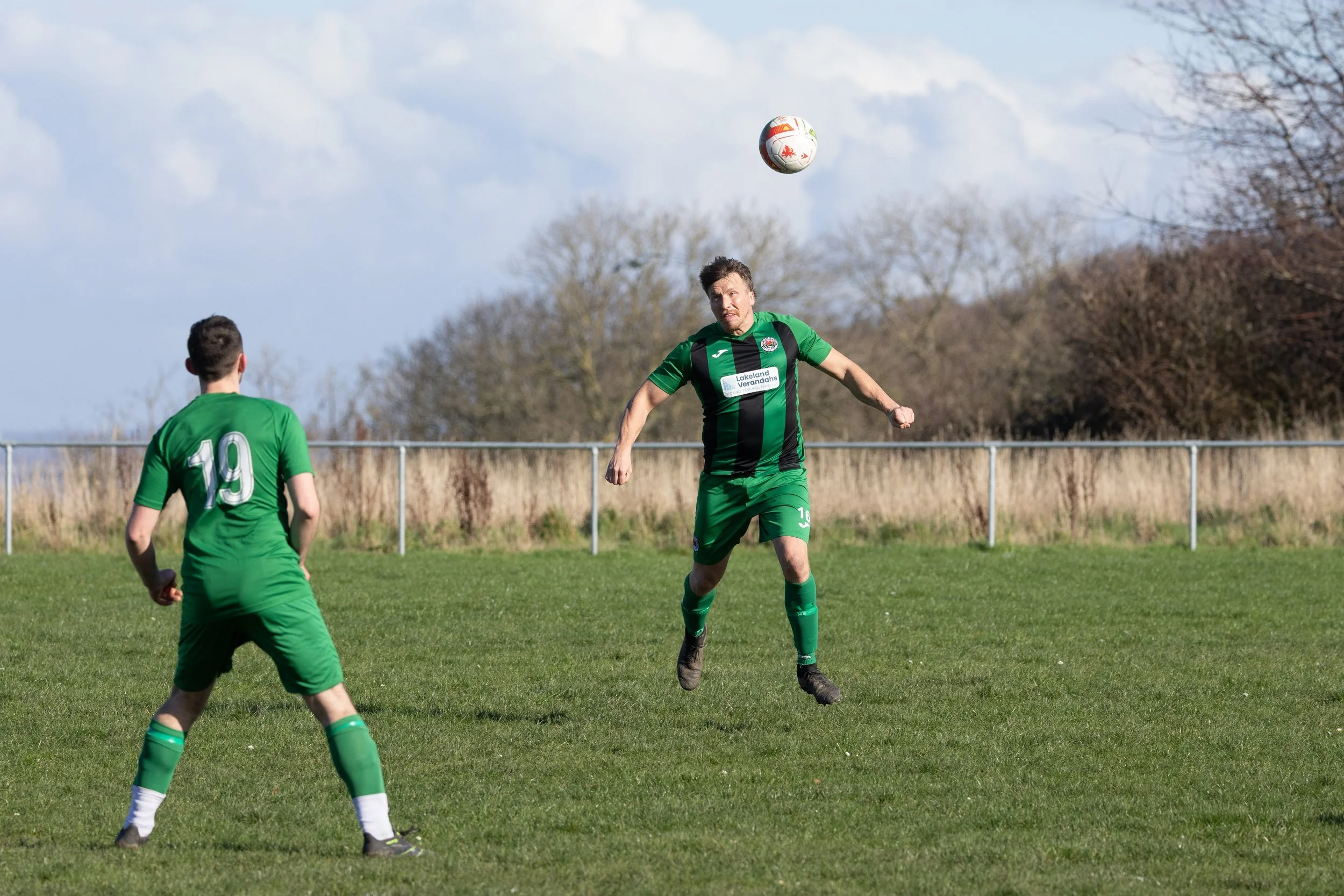 Two soccer players wearing green jerseys are on a grassy field, with one jumping to head the ball while the other watches. Trees and a metal fence are in the background under a partly cloudy sky.