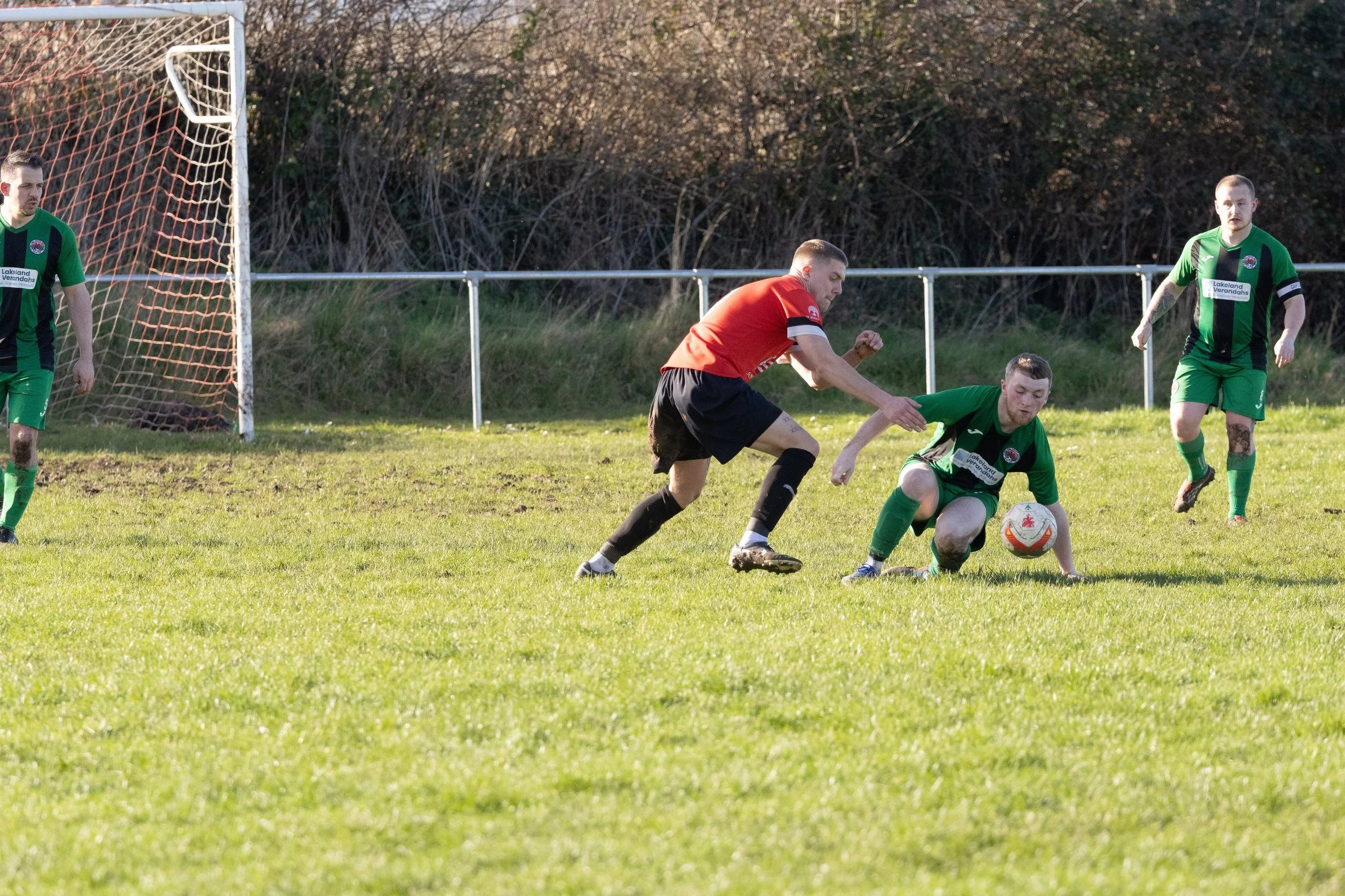 Soccer players in green and red jerseys during a game on a field with goalpost and fence in the background.