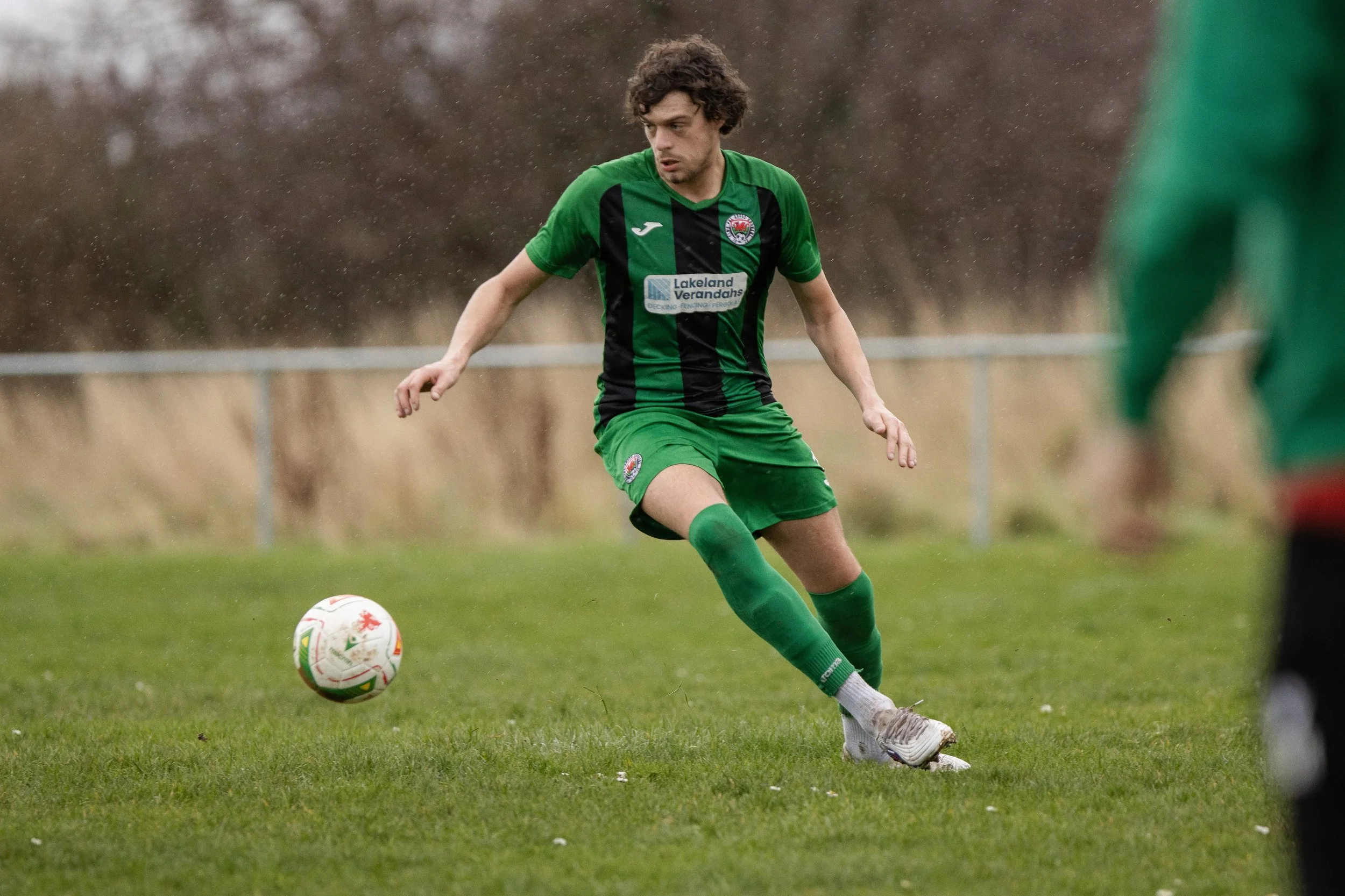 A soccer player in a green and black uniform kicks a soccer ball on a grassy field during rainy weather.
