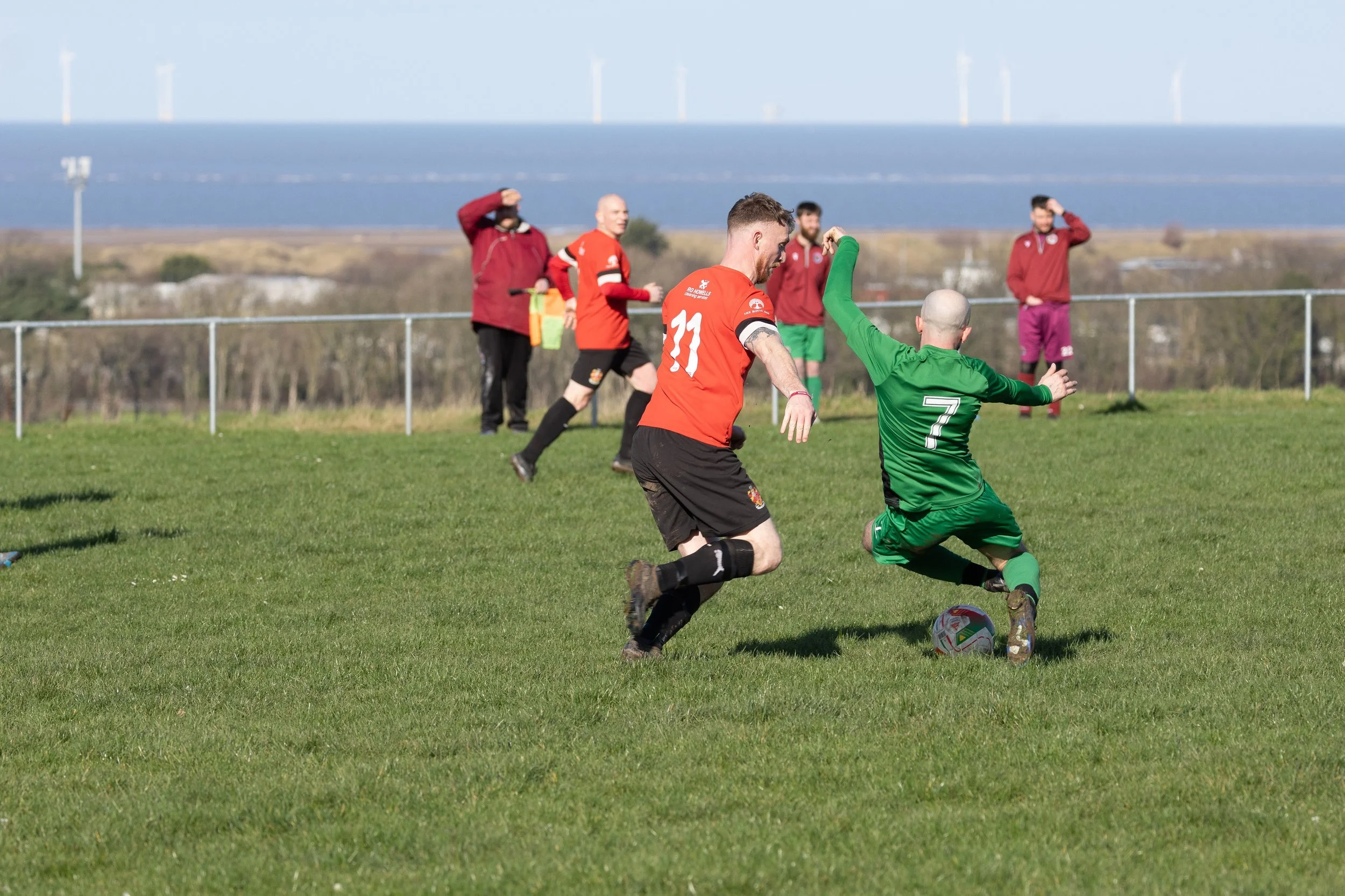Soccer players in red and green uniforms compete for the ball on a grassy field, with spectators in the background and a scenic landscape.